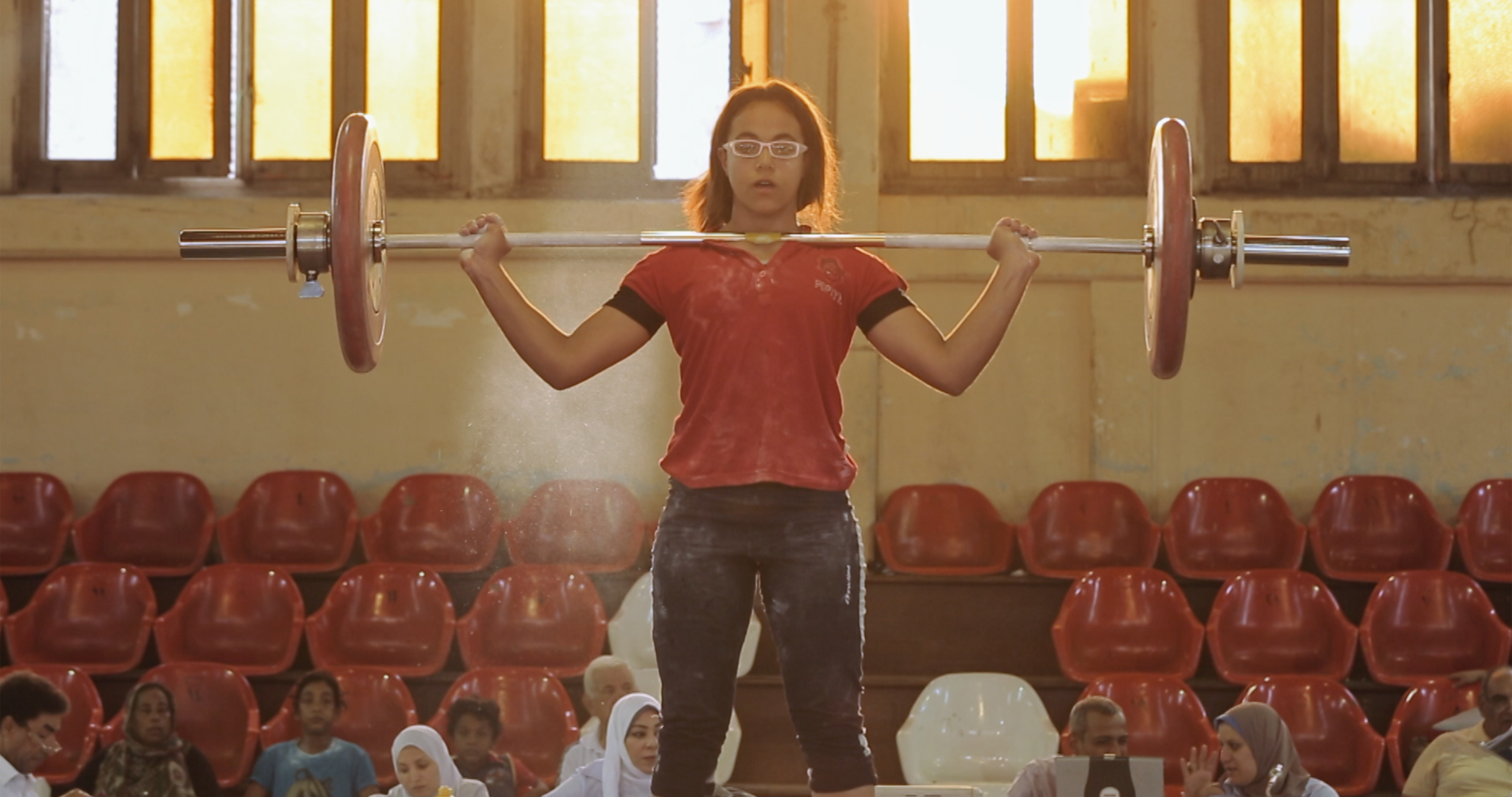 A girl in the process of weightlifting. Wearing white glasses and a red shirt, she holds the weighted bar on her chest. With red spectator seats in the background, the shot is illuminated by soft sunlight from the windows. 