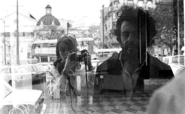 A woman and man are pictured in the reflection of a window. The woman is standing behind the man with a camera raised over her shoulder.