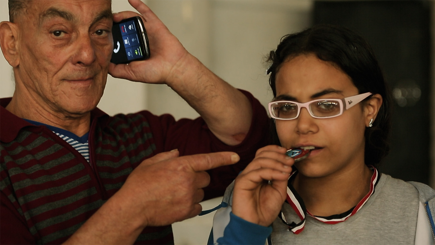 Close-up of a young girl with white glasses biting a medal. The arm of an older man points at the girl as he talks on his mobile phone. 