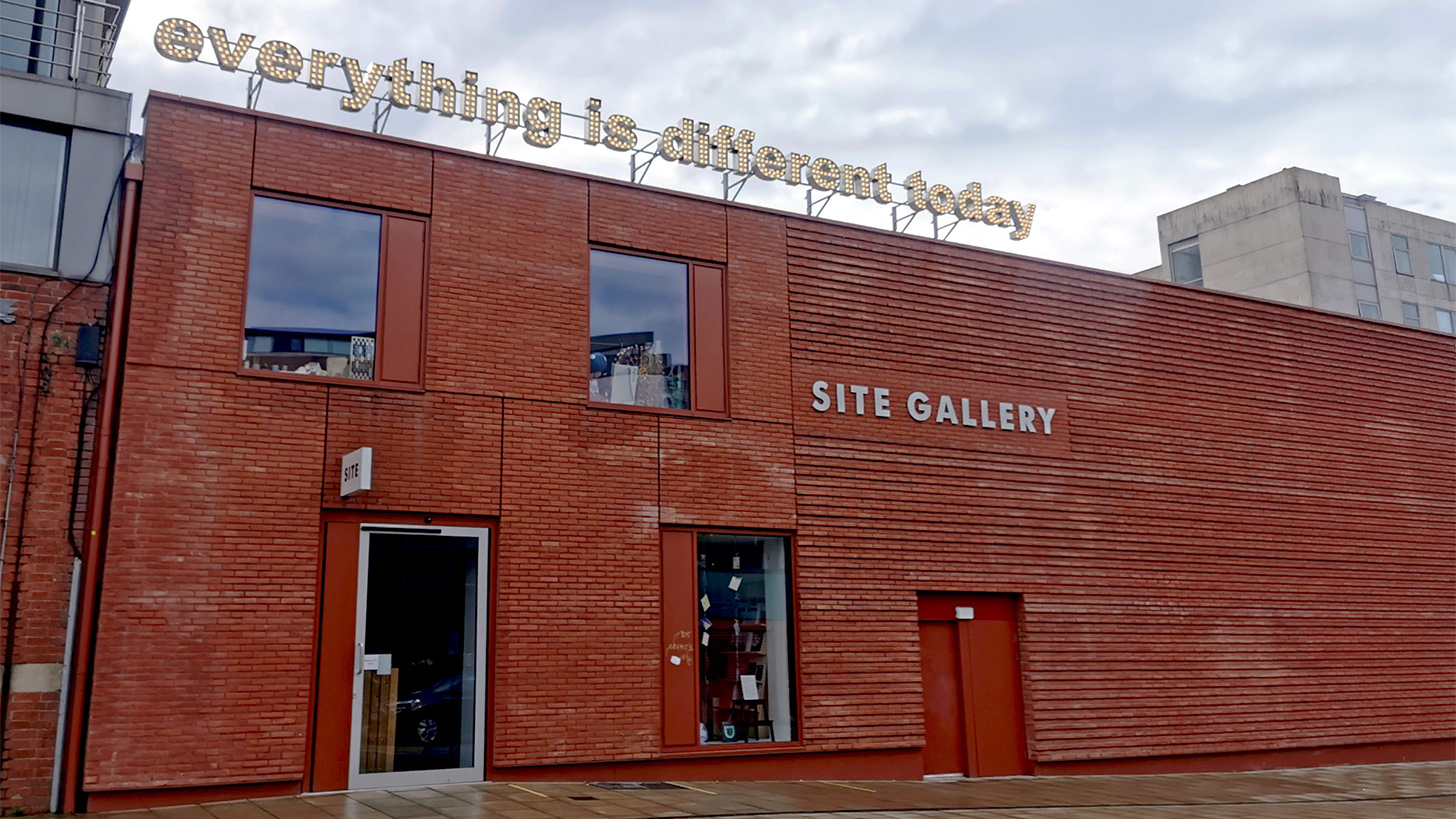 Sign reads 'everything is different today' in lights, sitting above a red brick building. White sign on the facade of the building reads'SITE GALLERY'