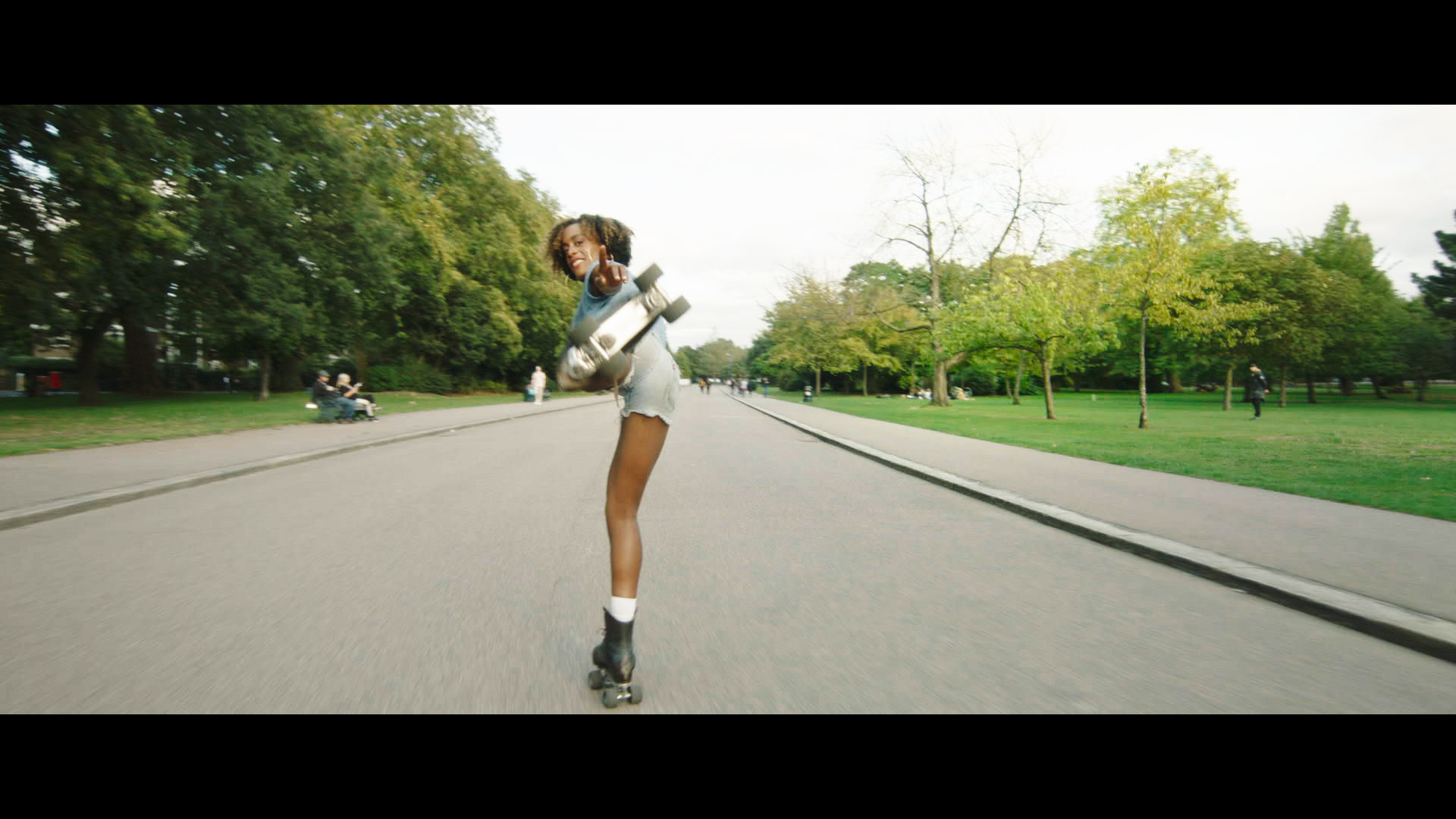 A woman rollerskates down a wide empty road. She turns her head and points with her left hand back to the camera, with her left leg outstretched behind her. 