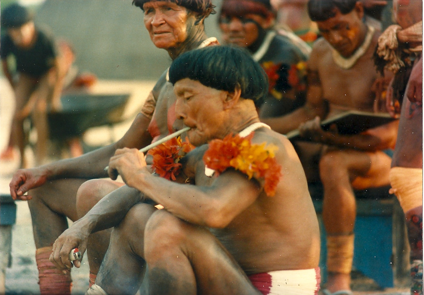 A group of indigenous people sat together. In the forefront a person is smoking a long cigarette.