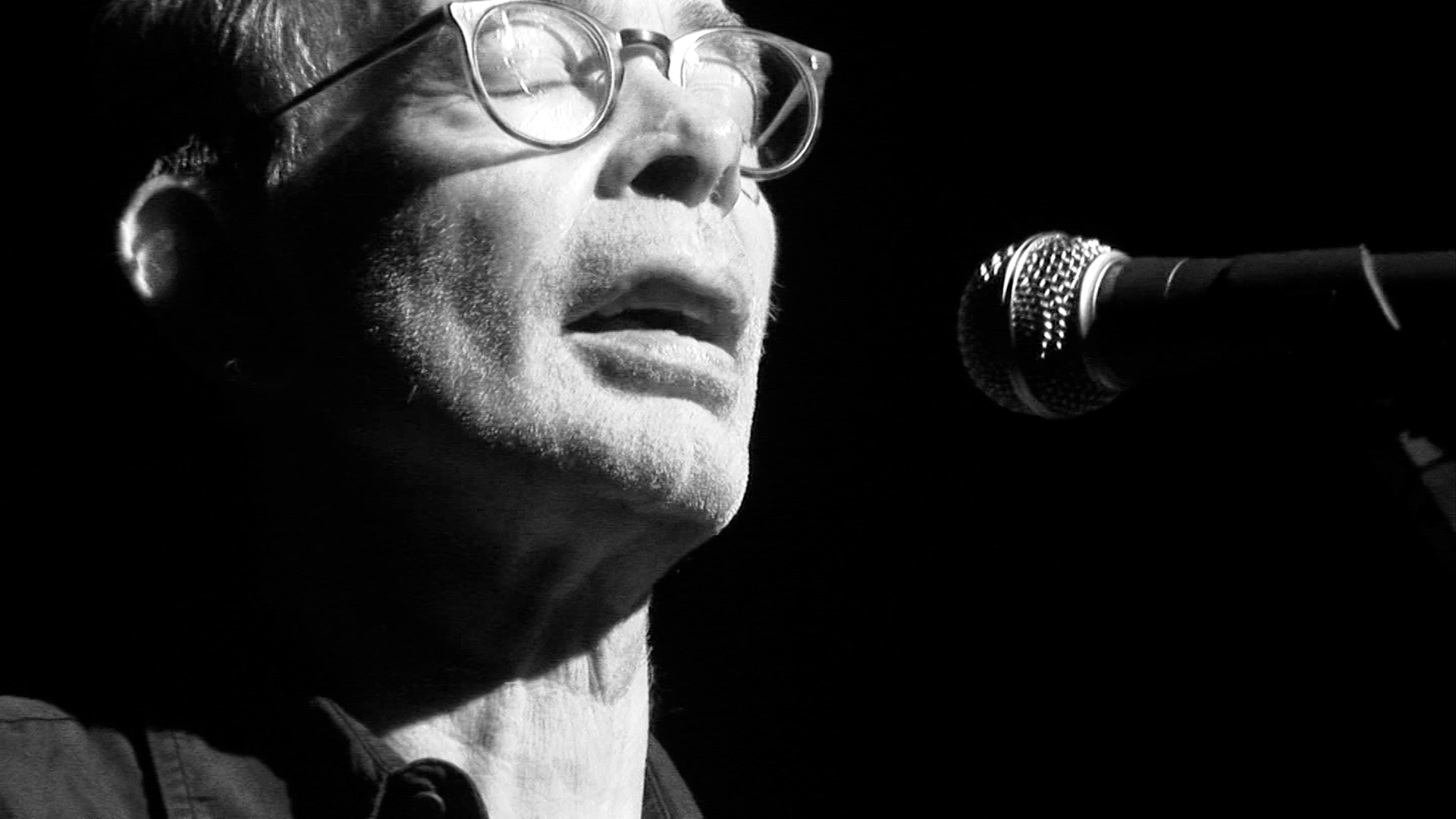 Black and white shot of a man in glasses singing into a microphone.