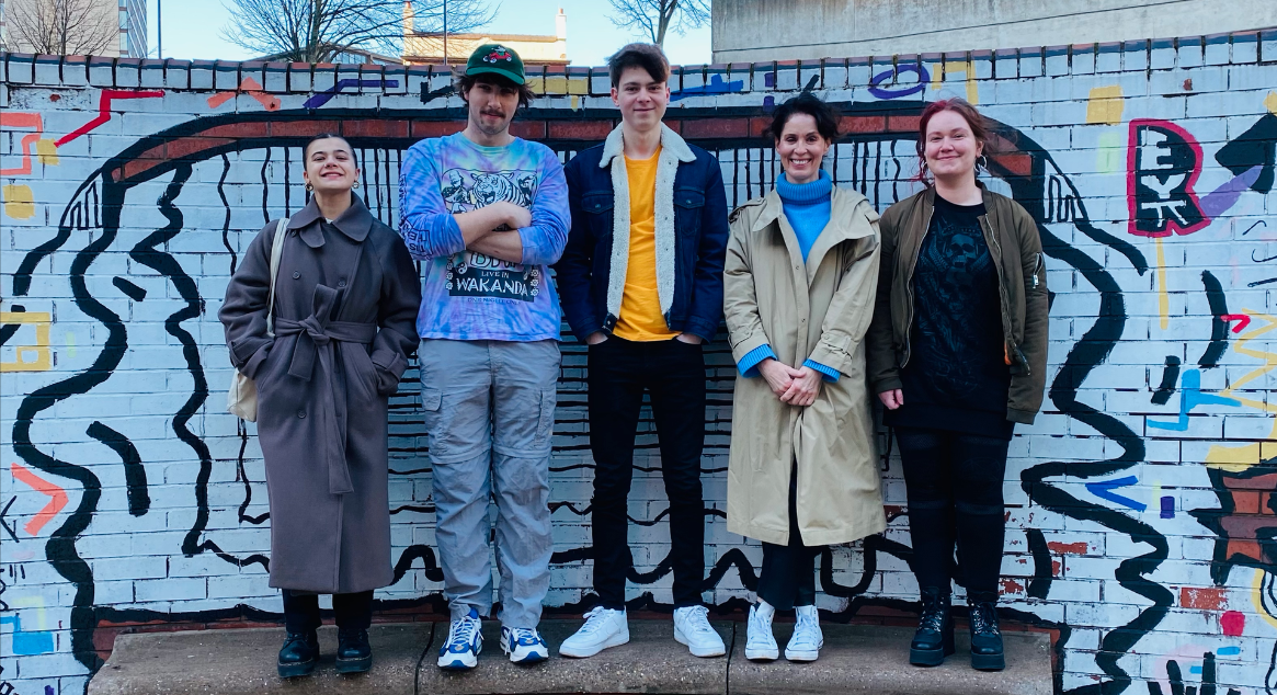 Five people stand side by side, in front of a colourfully painted brick wall. From left to right: Sophie Challis, Ralph Whittall, Brett Marsden, Sarah Curtis and Jess Campbell.