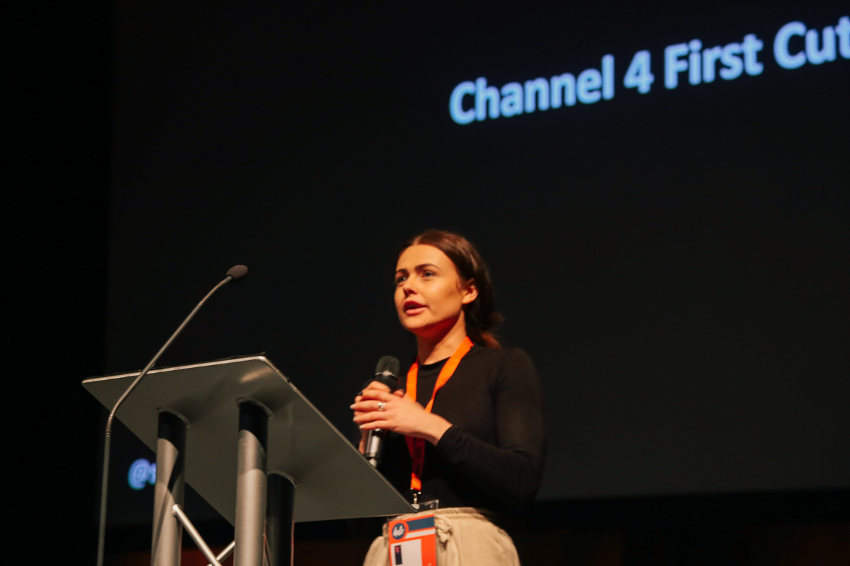 Woman standing behind a lectern holding a microphone. Behind her is a black screen with white text, reading 'Channel 4 First Cut'.