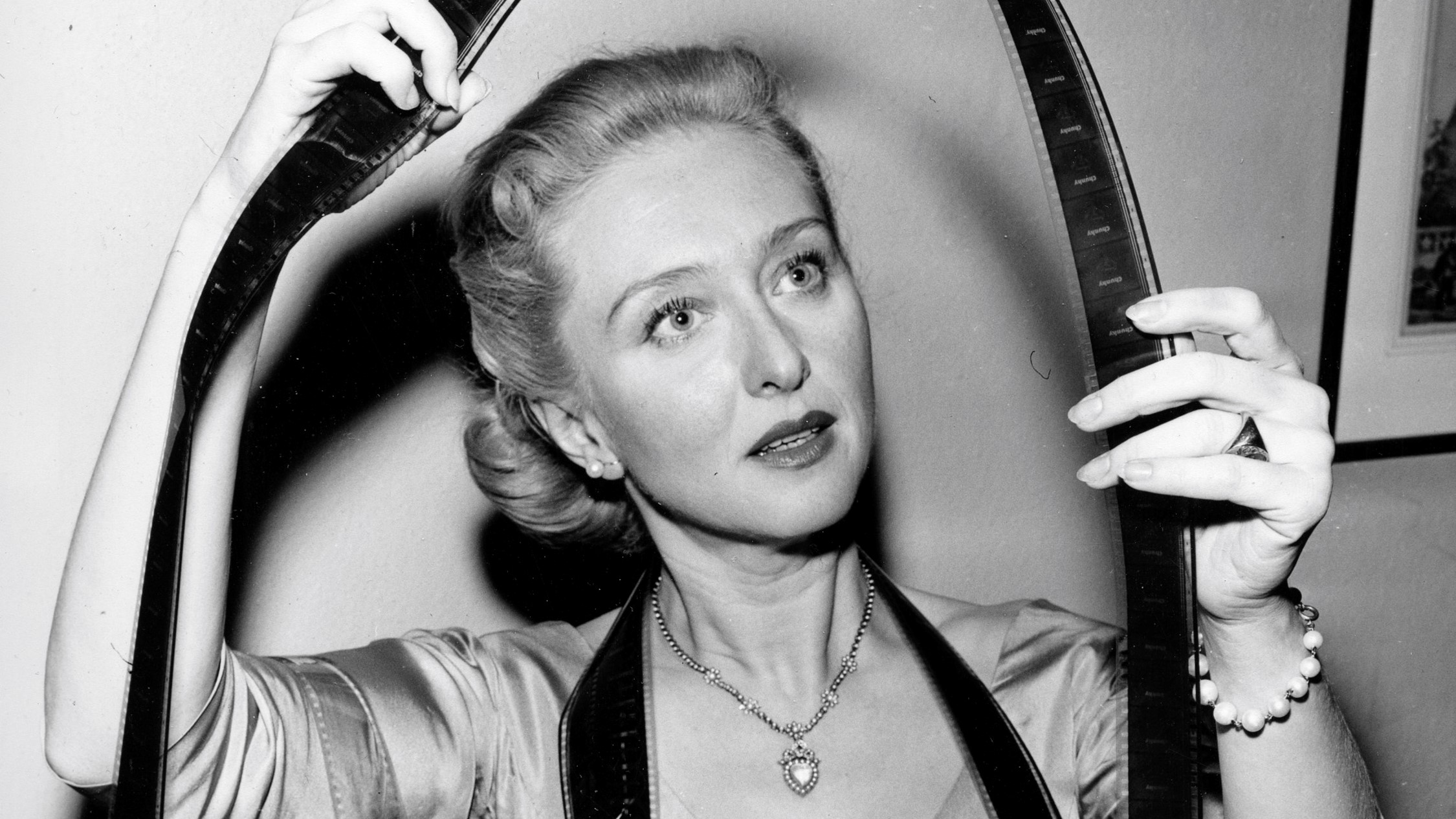 A black and white archival photo of a woman looking at a reel of film, which is held between both her hands and draped around her neck.