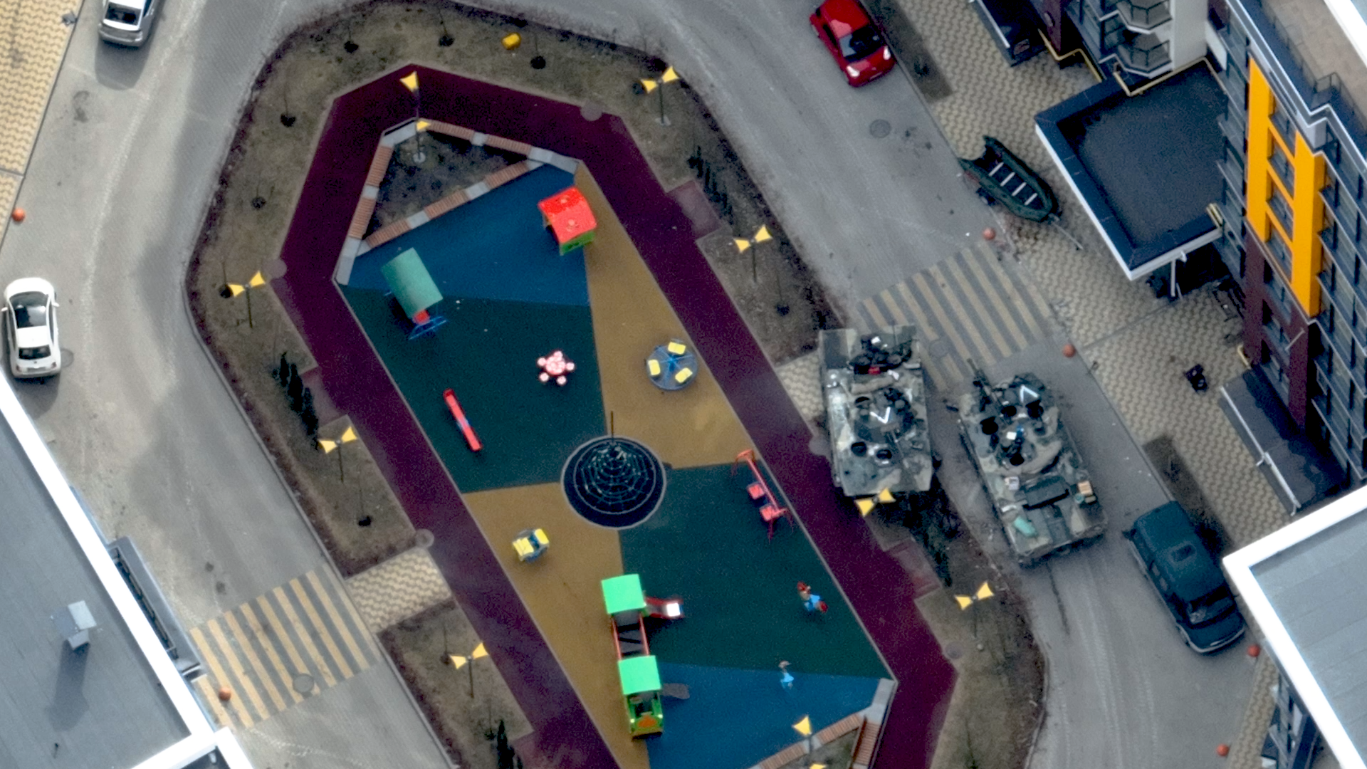 Still from One Day in Ukraine. Aerial view image of a children's playground, set in the middle of a roundabout. Beside the playground are parked cars, and two military tanks.
