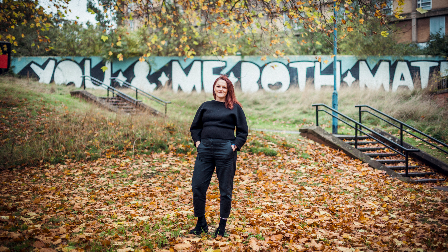 Annabel Grundy wearing a black jumper and trousers. Stood outside, surrounded by fallen autumn leaves. She's pictured against painted with a graffiti style mural - white text against a blue background reads: You and me both mate.