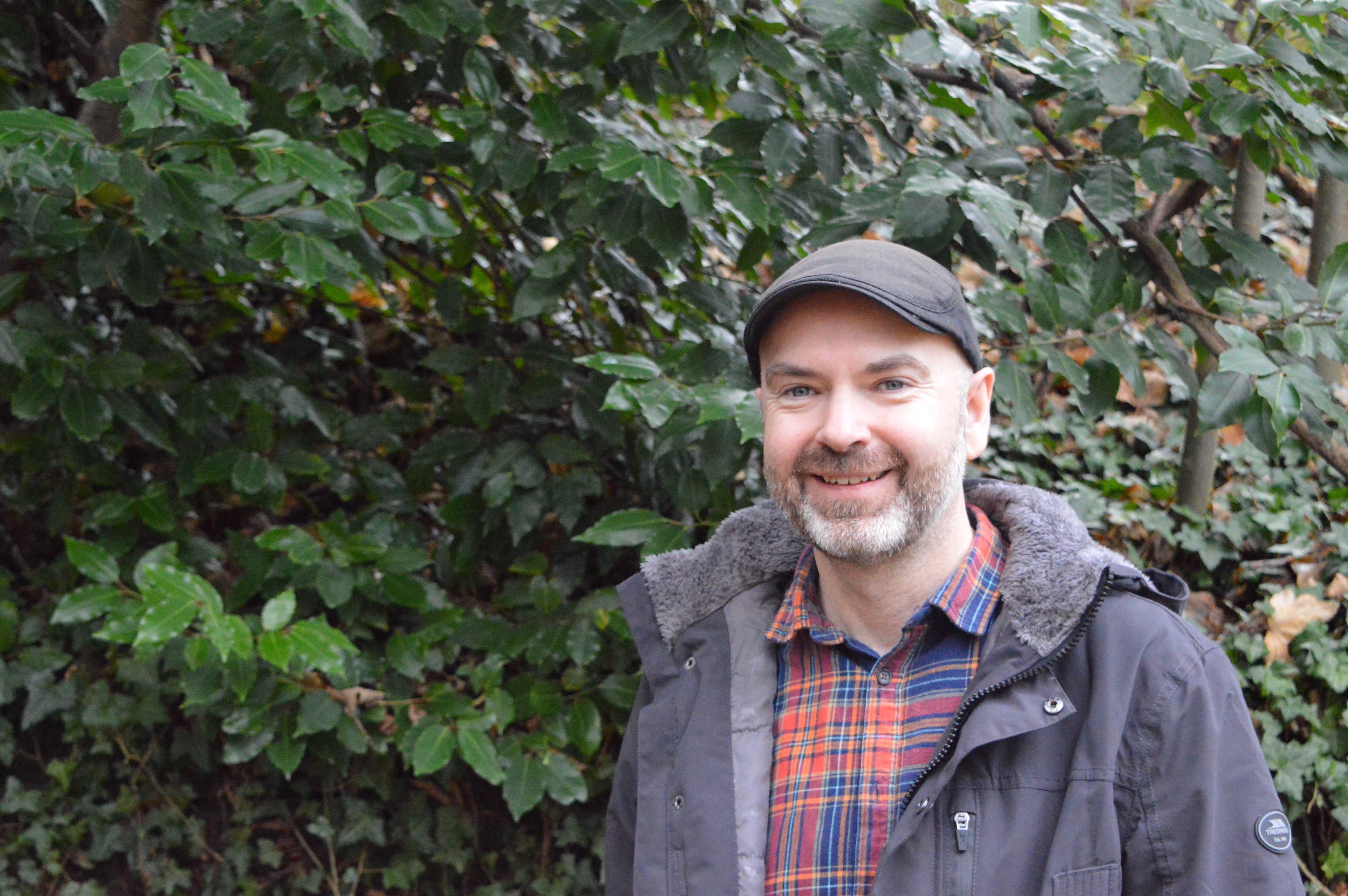 Pictured against a background of green foliage. Paul Steele standing to the right of the image, wearing a flat cap and winter coat.