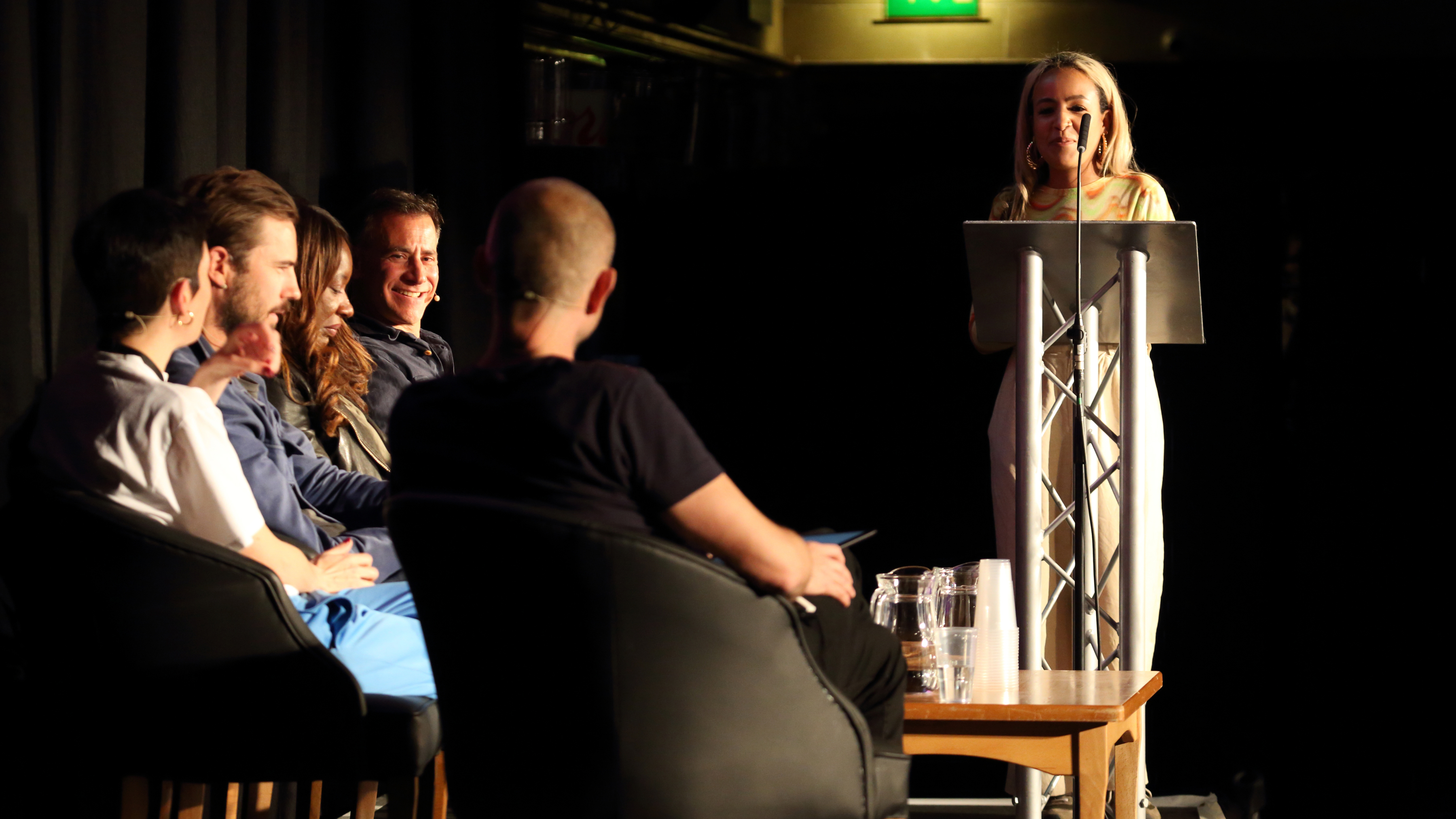 A woman with long, blonde hair is stood at a podium in front of a microphone. Beside her five people are sat on chairs, smiling.