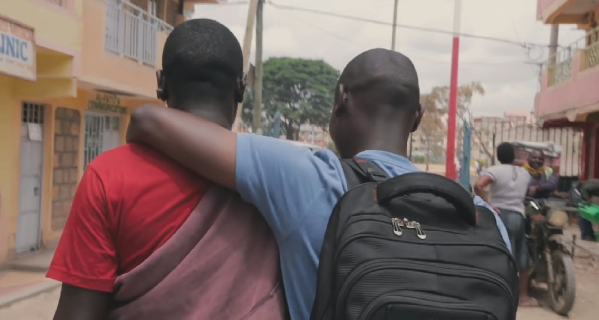 Image showing the back of two black men. One is wearing a blue t-shirt and a black backpack. He has his arm around the should of the other man, who is wearing a red t-shirt.