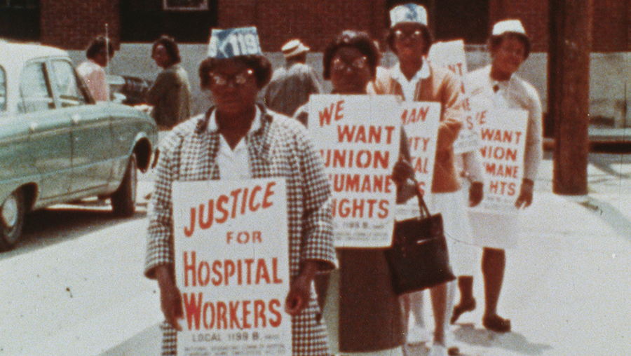An archival image of four black, female nurses. They're stood in a line holding protest signs.