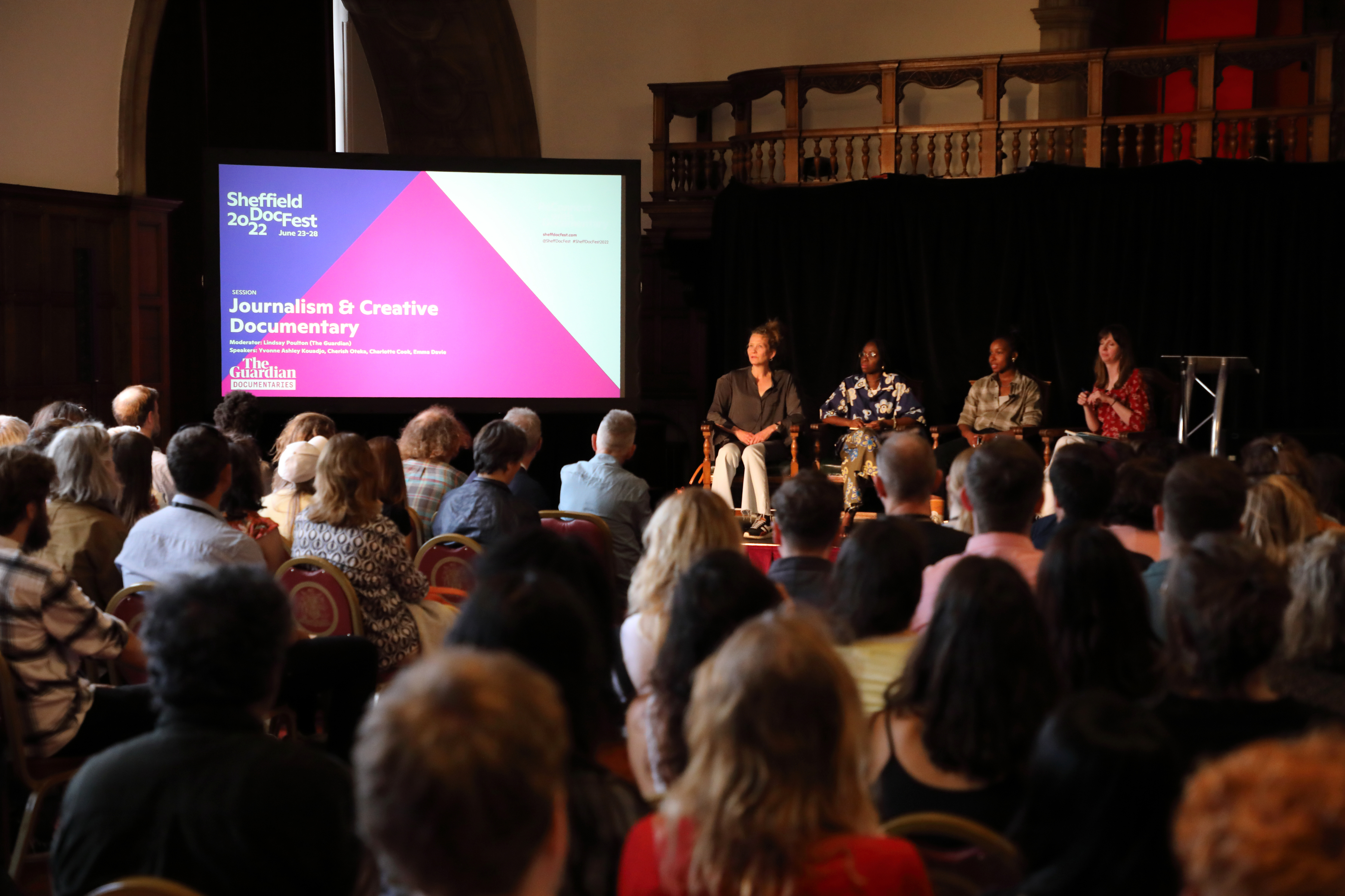 Image taken from the back of a full room showing people sat down, looking towards a stage where four panellists sit facing the crowd. To the left of the image is a large screen with a slide that reads: Journalism and Creative Documentary.