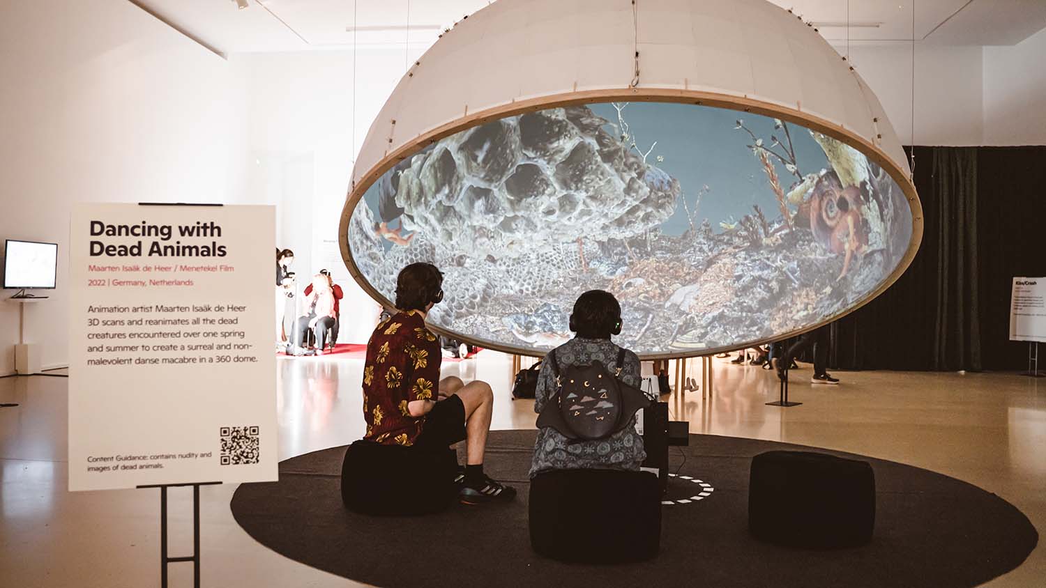 Image showing two people wearing headphones, sat on stools underneath a large half dome. Inside the dome, images of creatures, sea life and detritus is projected.