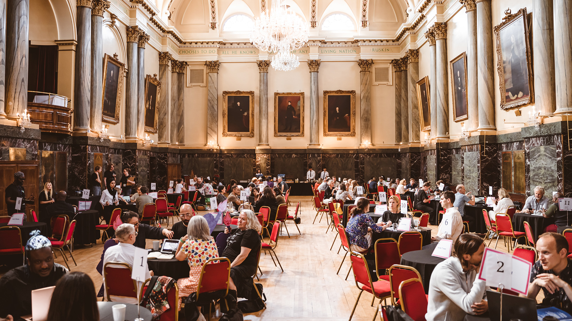 Groups of people chatting whilst sat around round tables, in a grand hall.