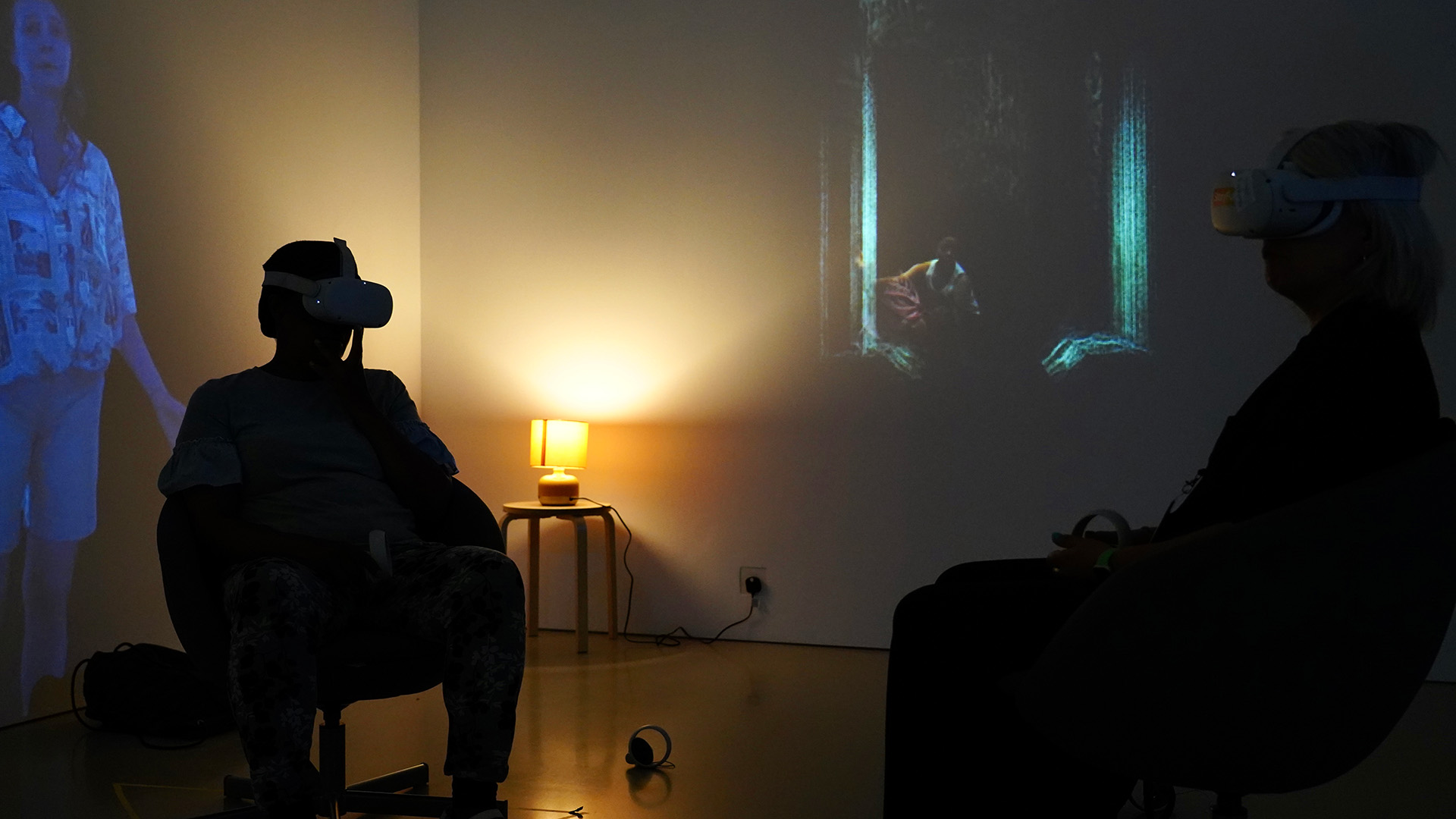 Two people sat down in a dark gallery space, both are wearing VR headsets.