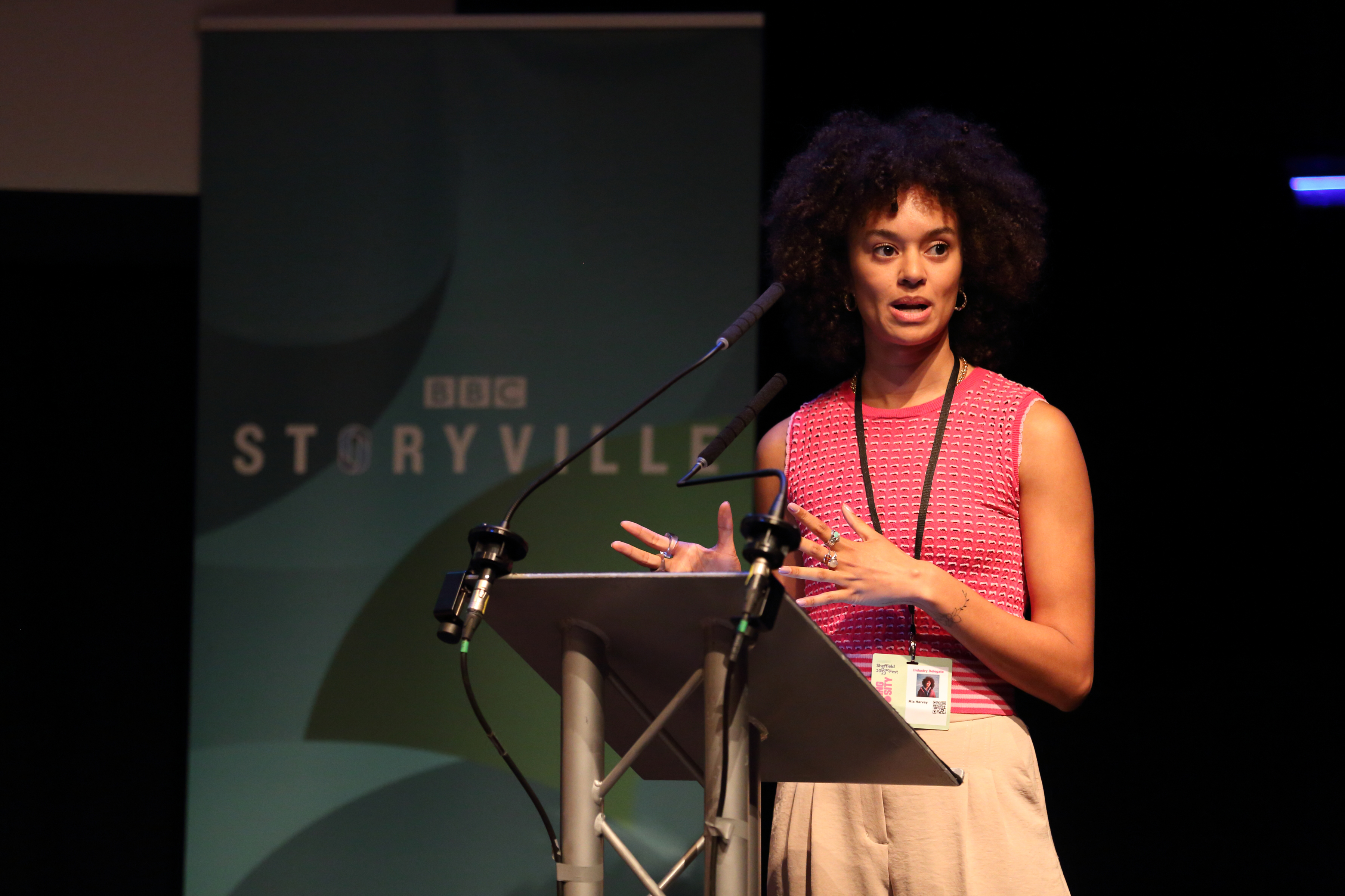 To the right of the image, a woman wearing a red top stands at a lectern with her hands waving in front of her expressively. Behind her is a pull up banner which says 'BBC Storyville'. 