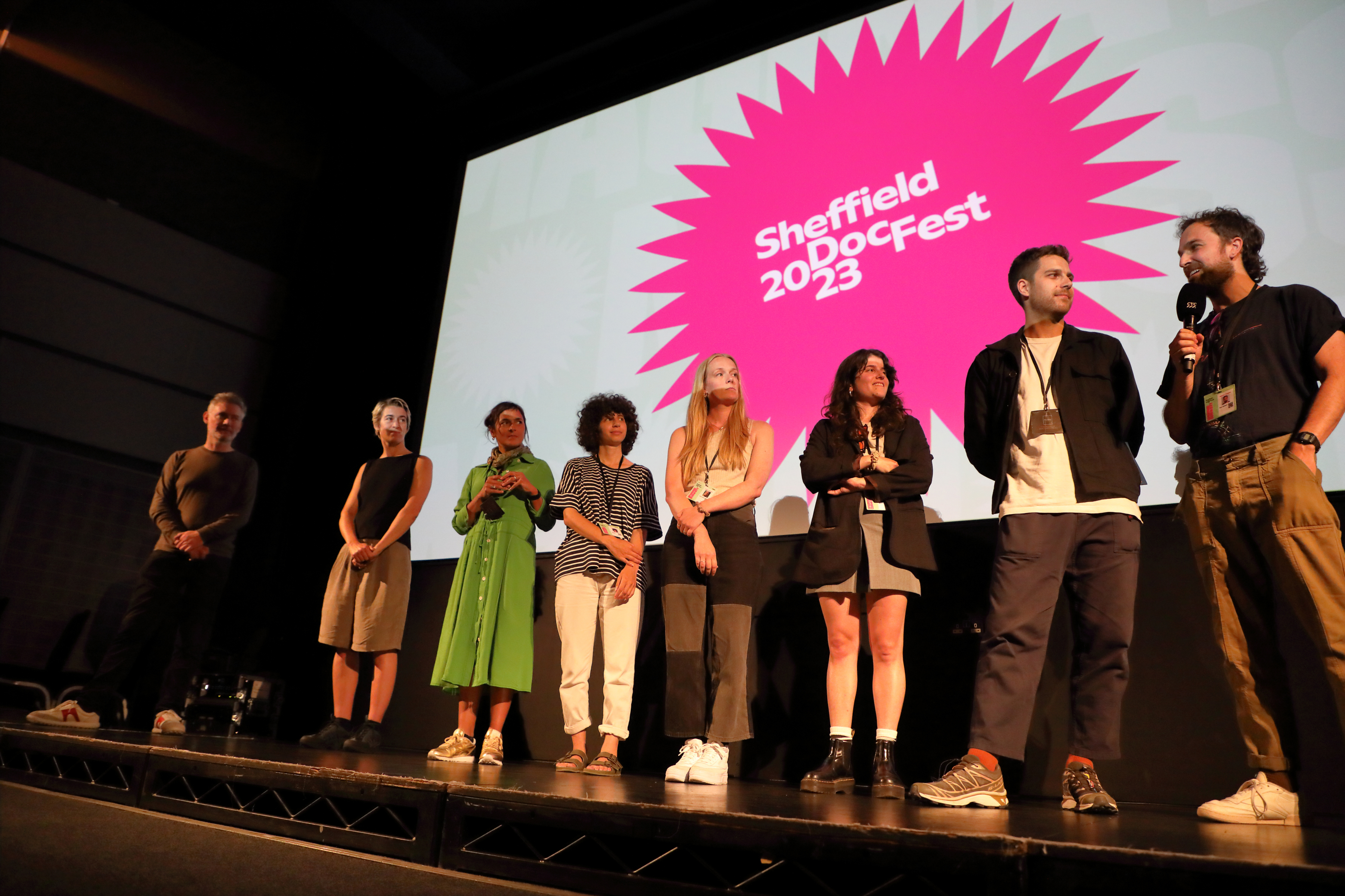 A group of people stand on a stage looking to the person on the far right who is holding a microphone. Behind them, a screen shows the words 'Sheffield DocFest 2023' 