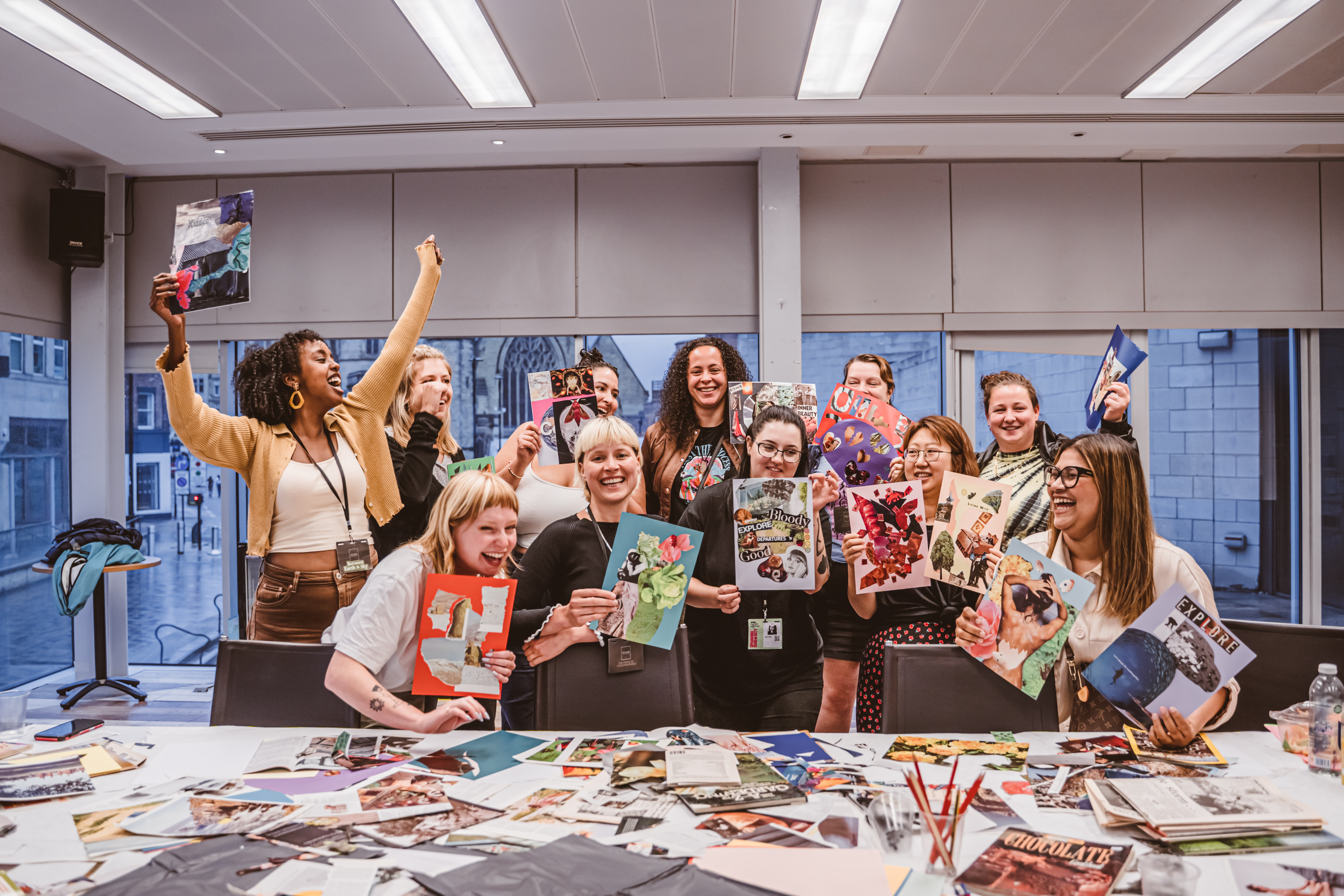 Group of people with wide smiles holding collages in the air with collage materials on a table in front of them