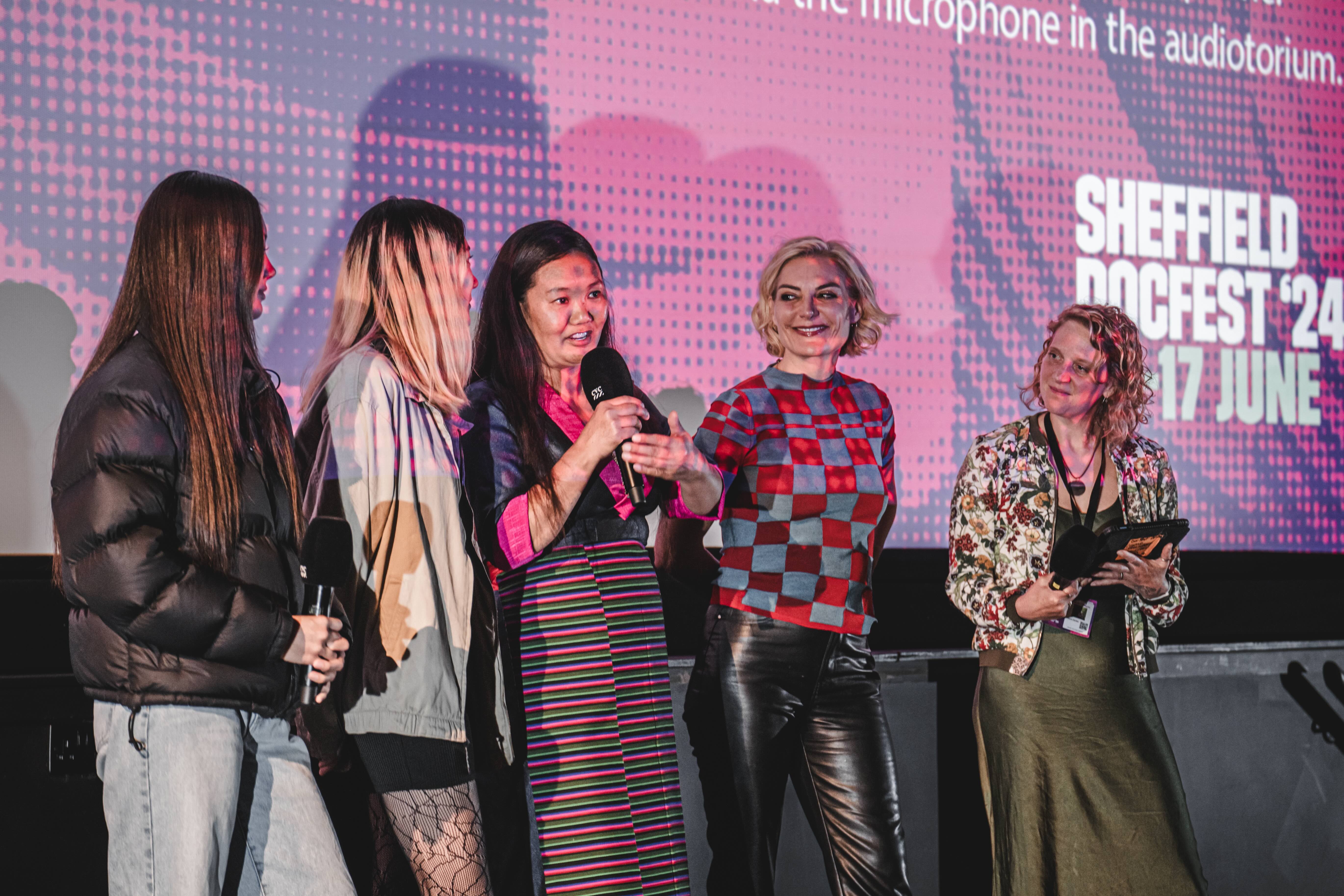 Five people stood on stage with a cinema screen behind them. The person in the middle holds a microphone to their mouth and is speaking into it. 