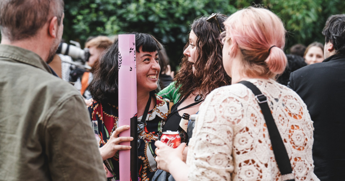 People surrounded by greenery at a busy drinks reception. In the centre of the photo two people looking at each other smile widely. The person on the left is holding a film poster rolled up in their hand.