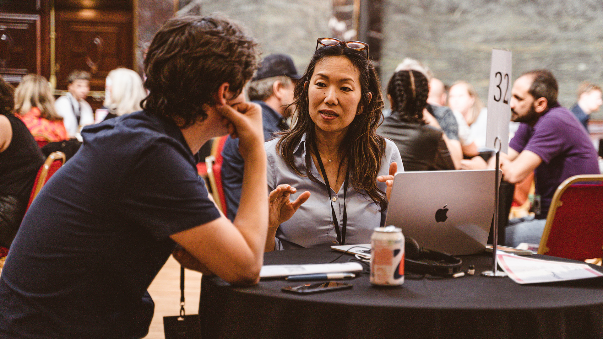 A woman and a man sat around a round table talking. The man has his back to the camera, the woman is facing the camera and has her glasses above her head and is gesturing with her hands.