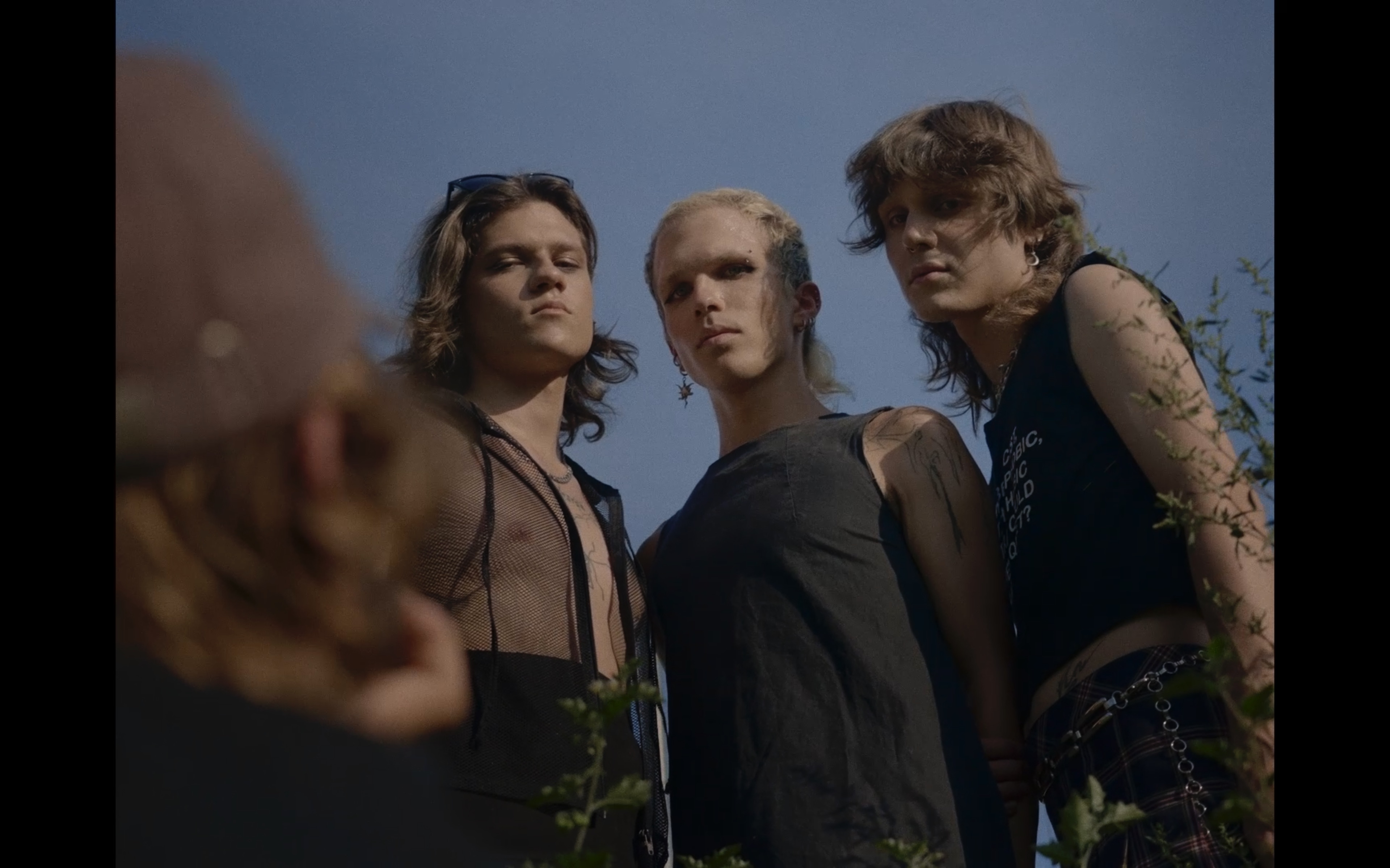 Three people stand close together, looking down towards the camera with serious expressions against a clear sky, partially framed by plants.