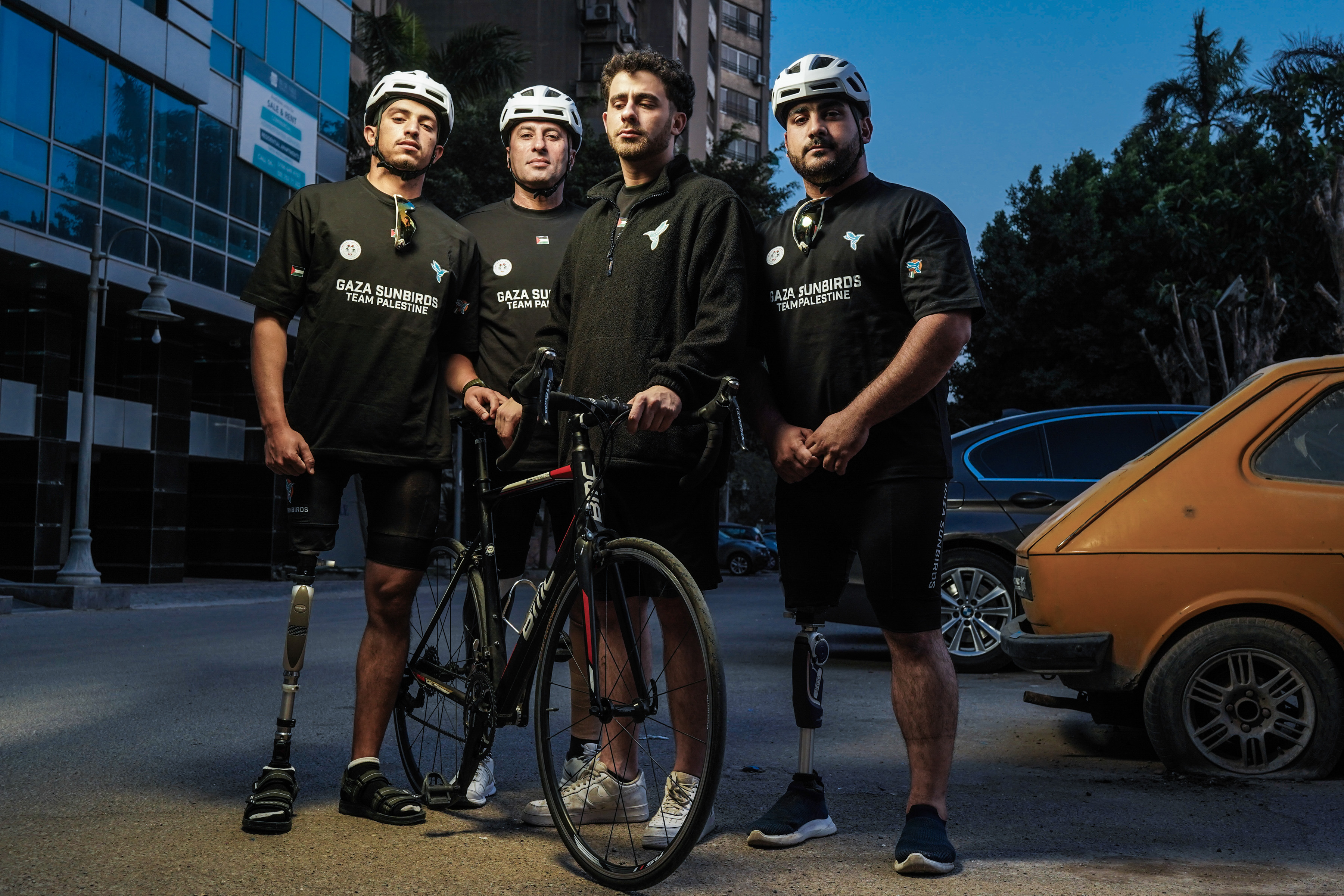 Four members of a team stand confidently with a bicycle in an urban street setting at dusk. Two of them wearing prosthetic legs, all dressed in team black shirts and three wearign helmets, illuminated by dramatic lighting.