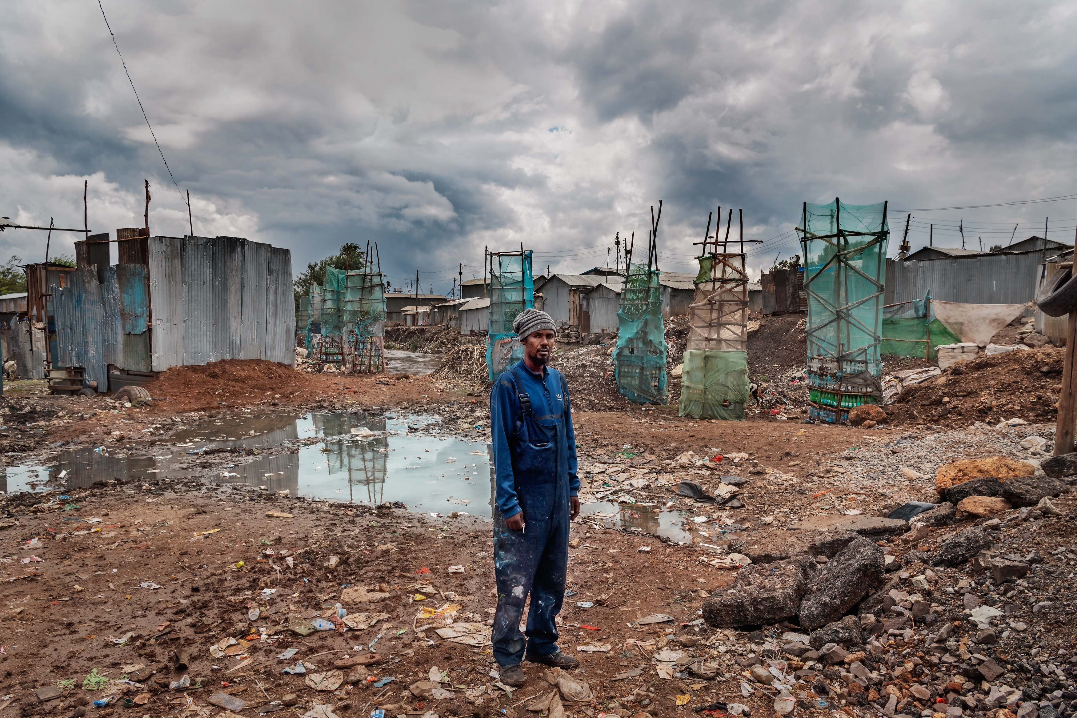 A black man in a paint-stained blue jumpsuit and striped beanie stands amid a muddy, debris-strewn construction site. Behind him, skeletal building frames covered in green netting rise against a backdrop of dark, stormy clouds.
