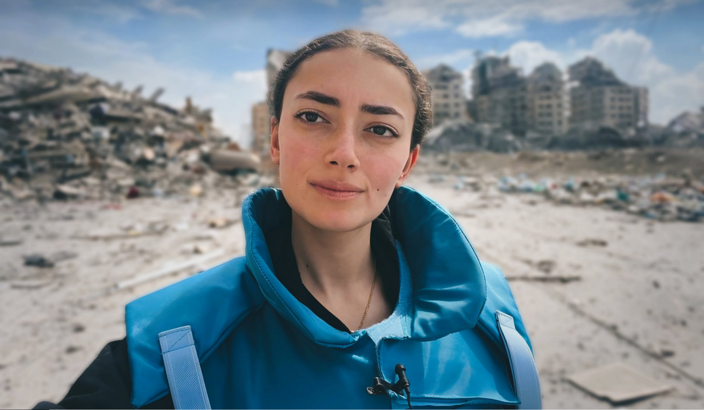 A woman wearing a bright blue protective vest stands in the foreground, looking directly at the camera. Behind her is a scene of destruction: large piles of rubble and partially collapsed buildings under a partly cloudy sky. The setting appears to be a disaster or conflict zone.
