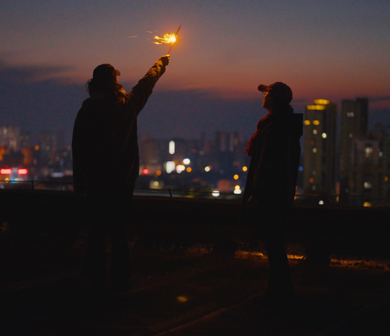Two figures standing on a rooftop at dusk or early night, with a cityscape glowing in the background. One person is holding a lit sparkler aloft, casting a warm light and small sparks into the air. The silhouettes of both individuals are visible against the fading light of the sky.