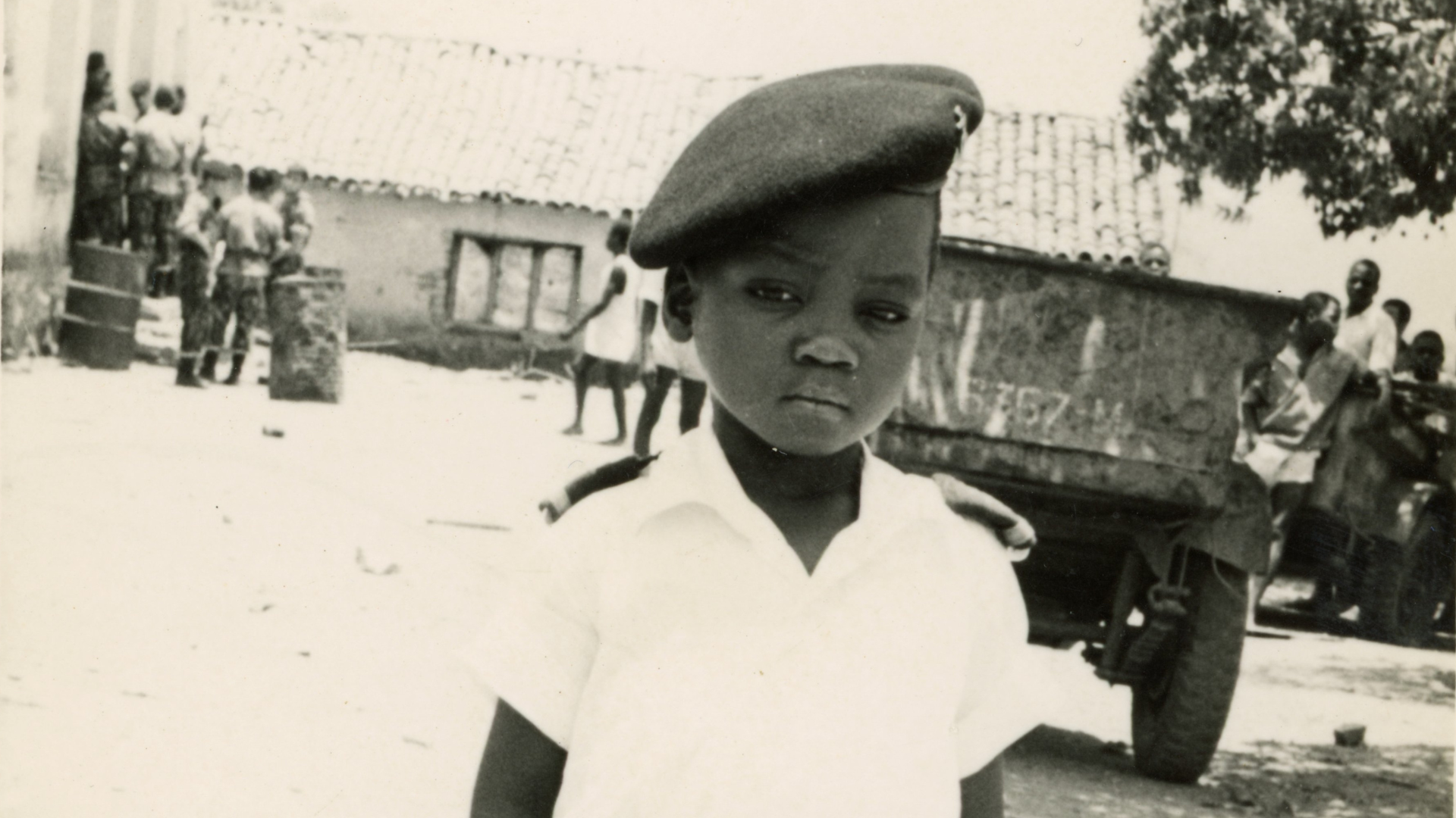 A black-and-white photograph, where a young boy wearing a military-style beret and white shirt stands solemnly in the foreground. His expression is serious and composed beyond his years. Behind him, a group of soldiers and civilians gather near a weathered building and an old military vehicle.