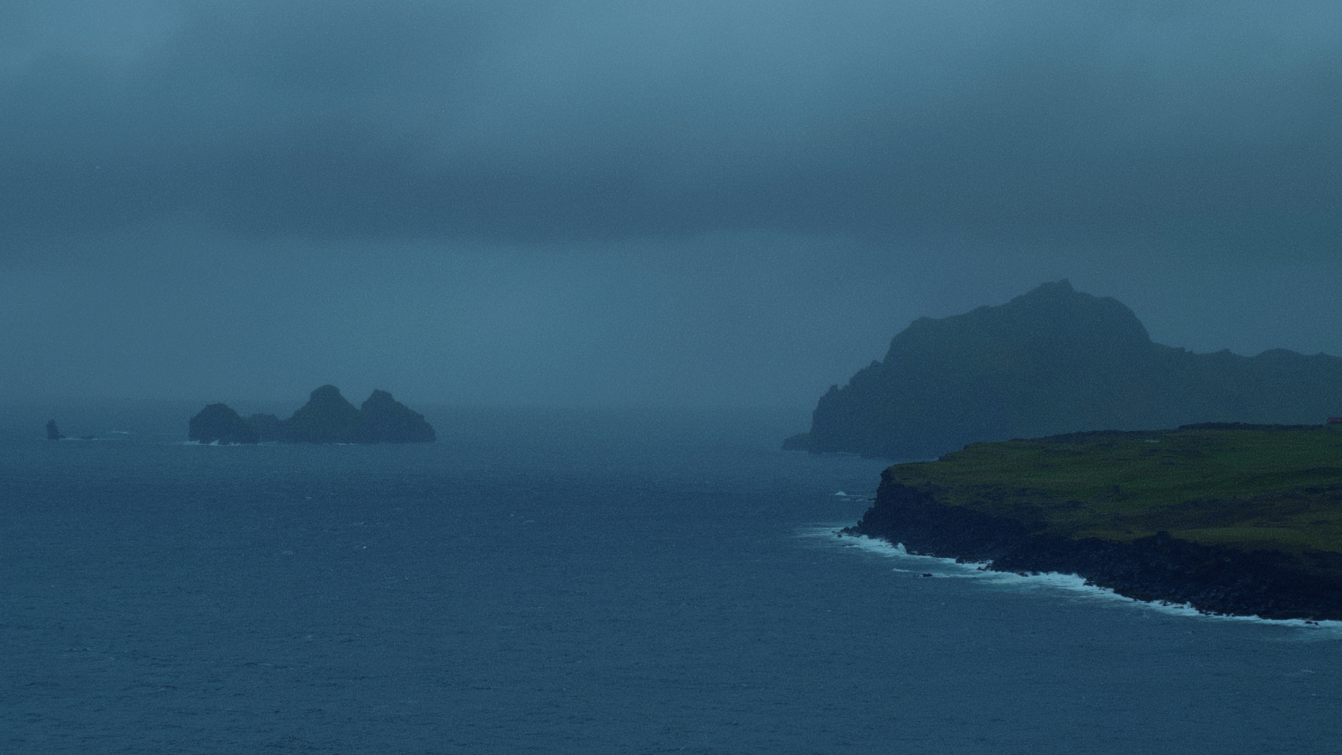 A brooding coastal scene under overcast skies, where stark cliffs stretch into the sea from the right, their dark rock faces contrasting with a narrow strip of green grass above. In the distance, rugged islets rise from the water, shrouded in mist and shadow.