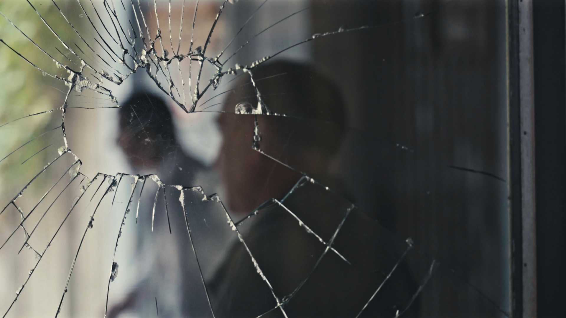 A cracked windowpane through which we glimpse a blurred human figure in the background. The broken glass radiates outward from a central impact point, creating a spiderweb-like pattern that visually fragments the person beyond.