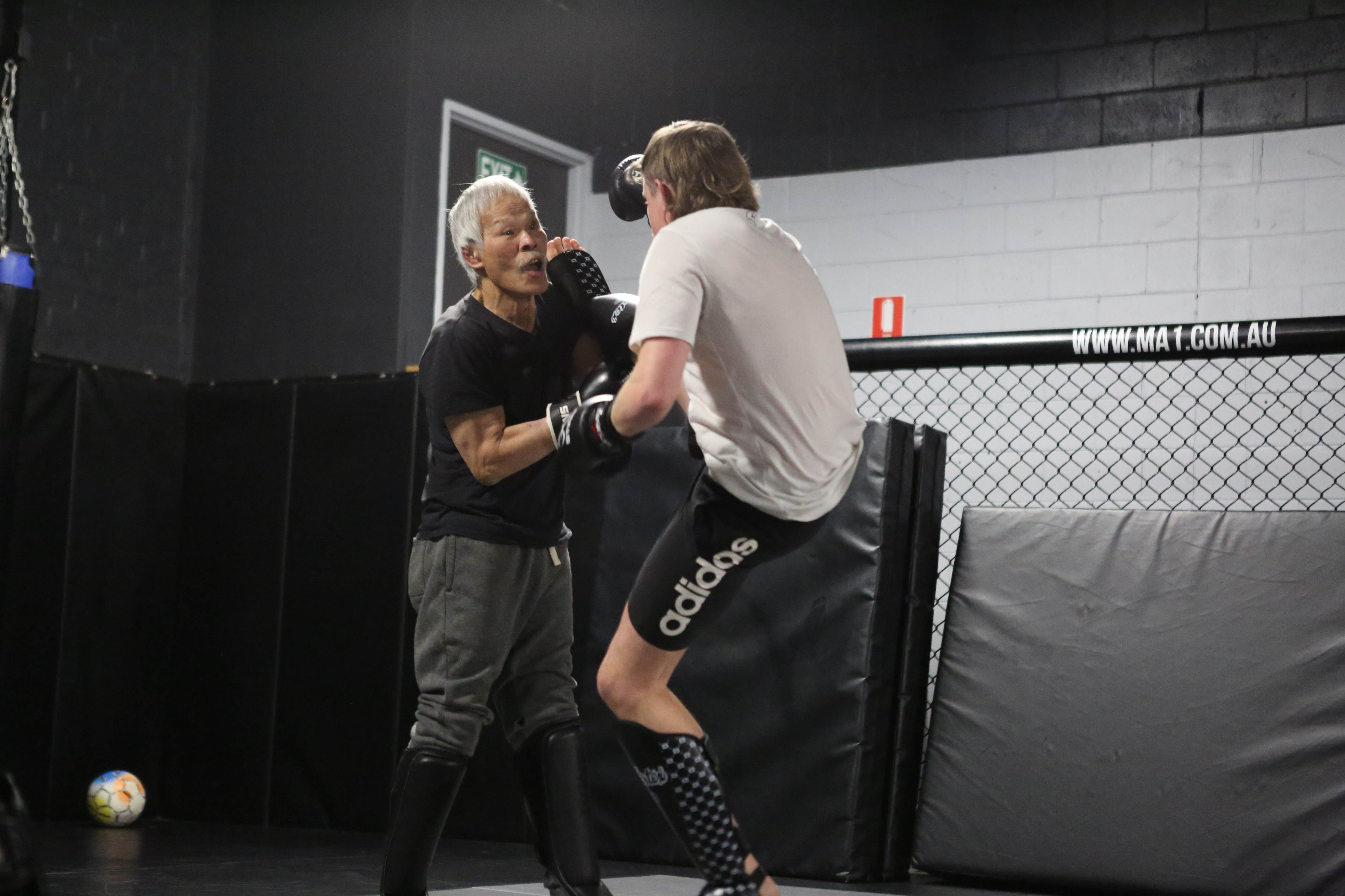 Two people are sparring or training in a martial arts gym. One older Asian person with grey hair is holding focus mitts while the younger person in gloves and shin guards is practicing strikes. They are inside a caged training area with padded walls and a soccer ball in the corner.