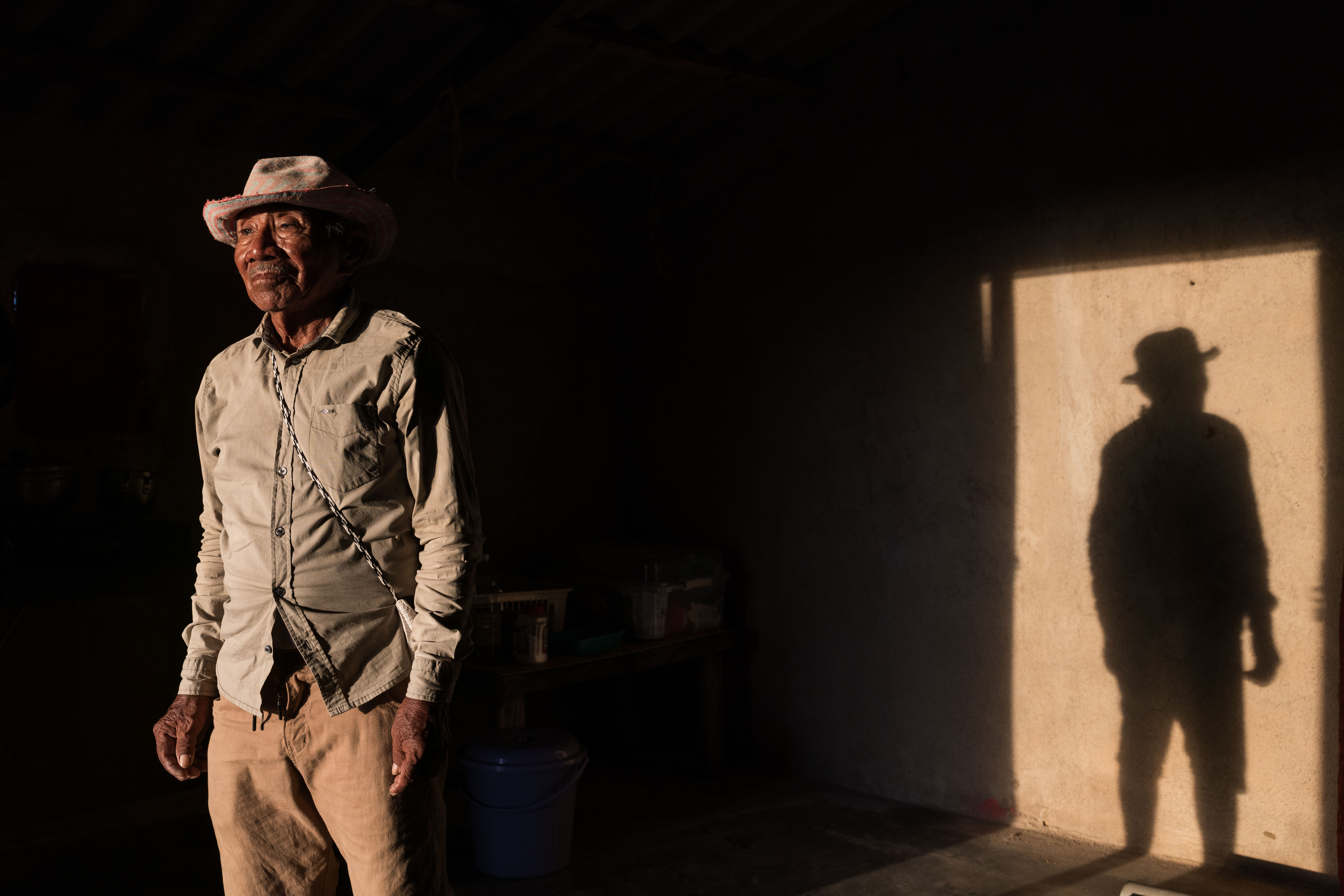 An older man standing in a dimly lit room, with strong light casting a long, defined shadow of him on the wall. He wears a hat and simple clothing, with a contemplative or slightly solemn expression.