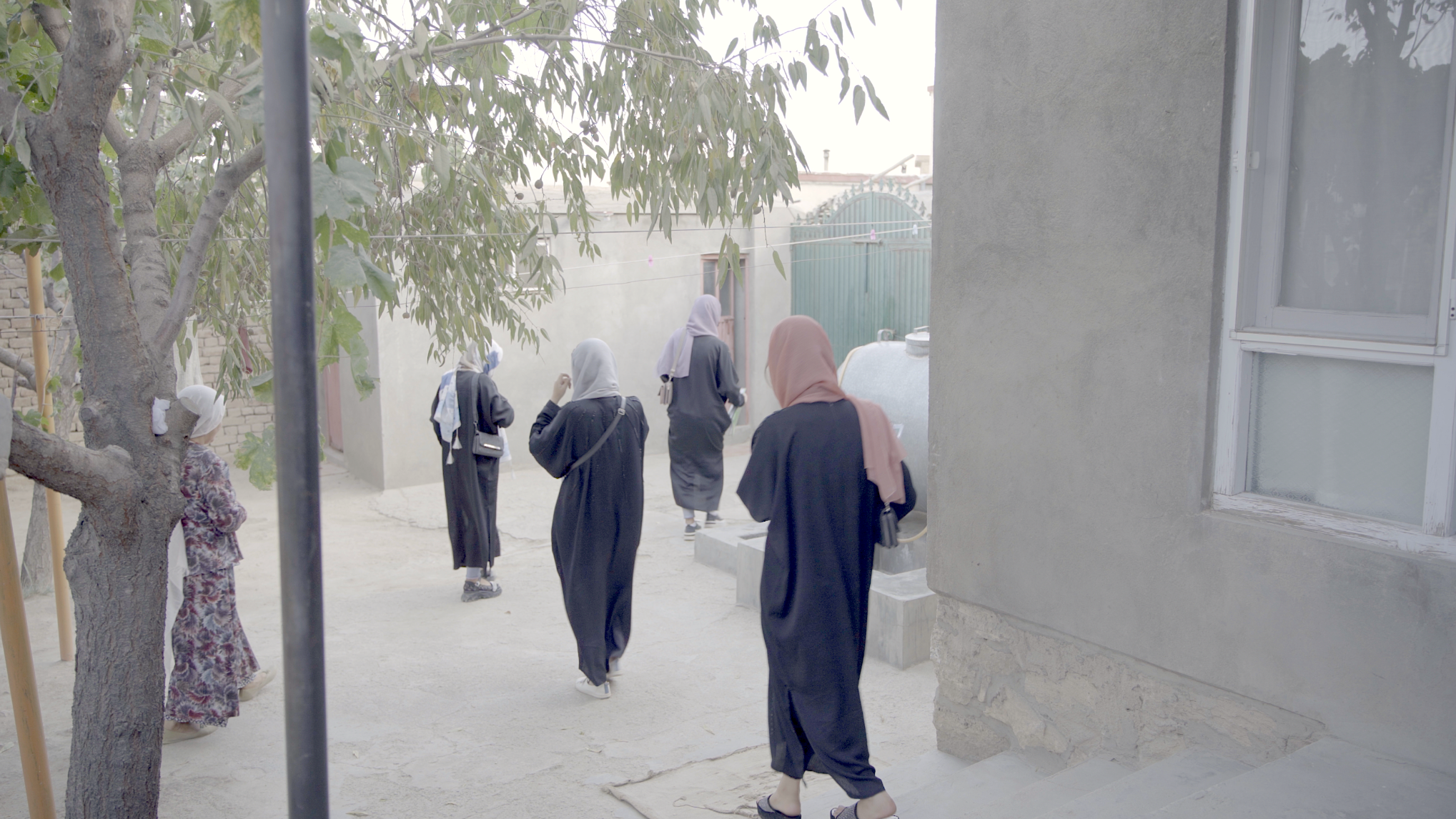 A group of women or girls, all wearing long black clothing and headscarves in various colors, are walking away from the camera in a concrete courtyard. There is a tree to the left, a grey building with a window to the right, and a green gate in the background. One woman is wearing a patterned dress and headscarf and stands near the tree, facing slightly away. The scene is softly lit, with an overcast or diffused daylight.