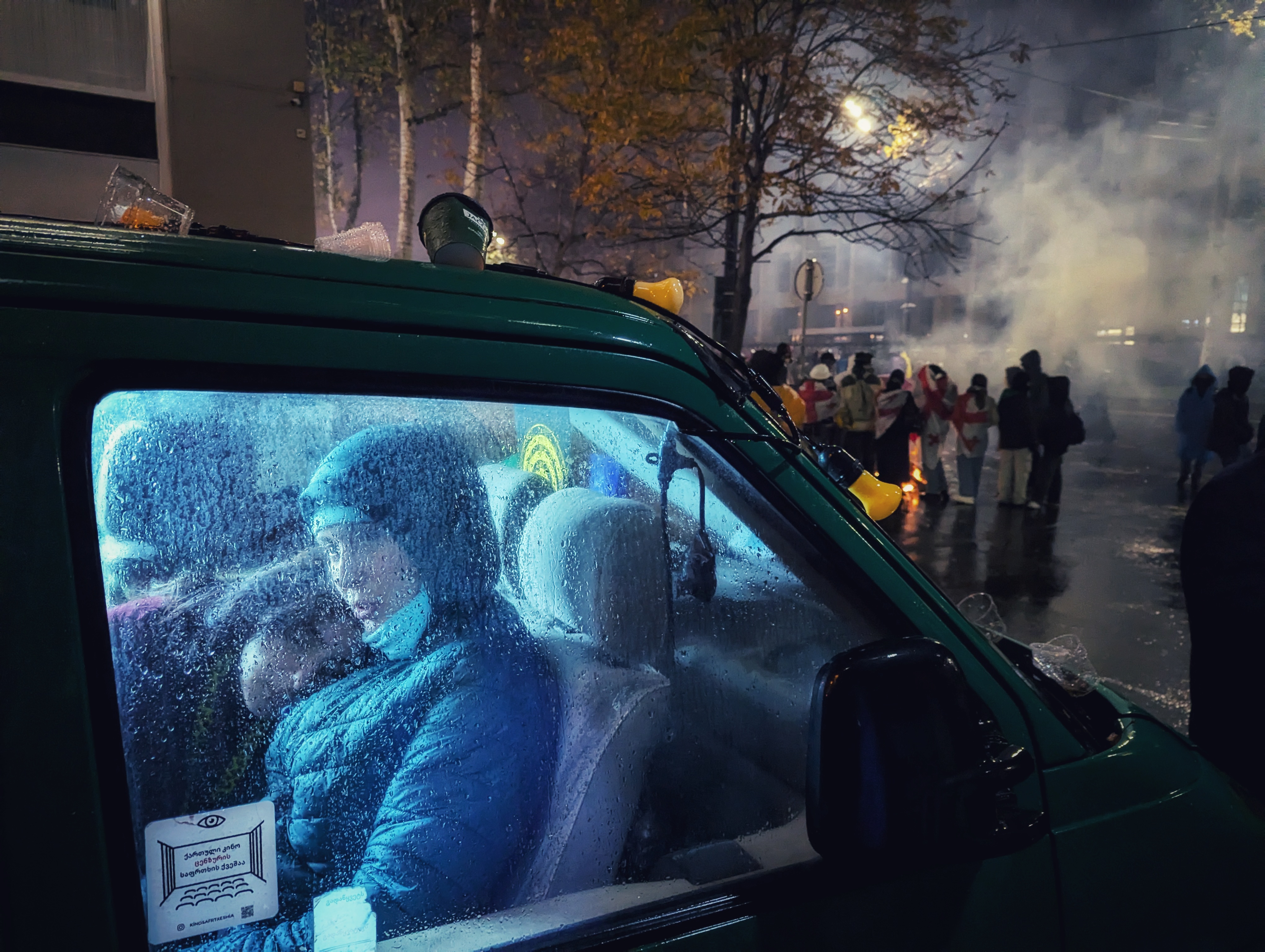 Inside the rain-specked window of a vehicle, a woman cradles a child, their peaceful moment lit by cold blue light. Outside, smoke, fire, and a crowd in protest or crisis under streetlights and bare autumn trees.