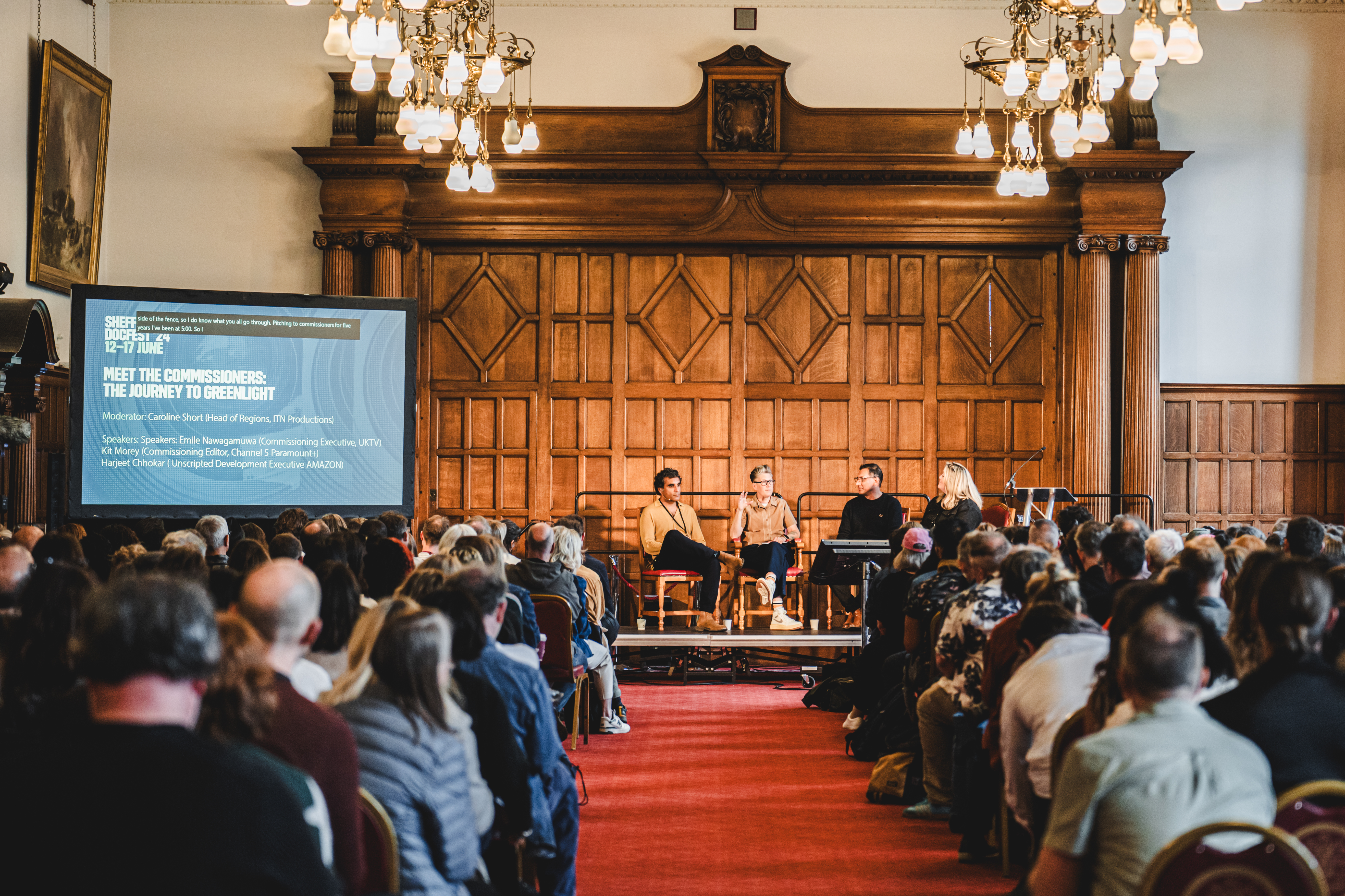 A large hall of people facing towards a group of people sat on a stage with a large wood paneled wall behind them.