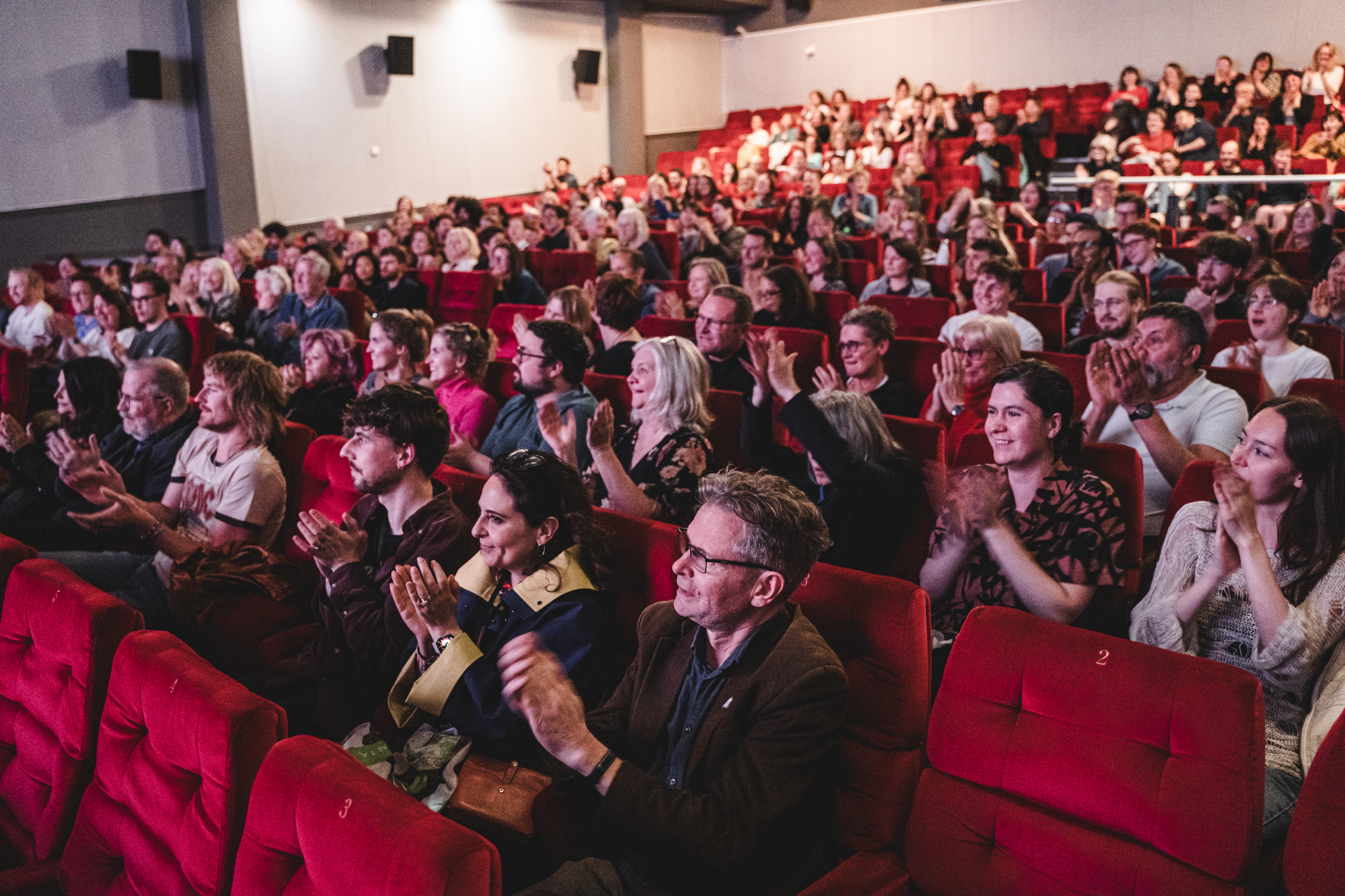 A busy cinema crowd sat on red velvet seats applauds.