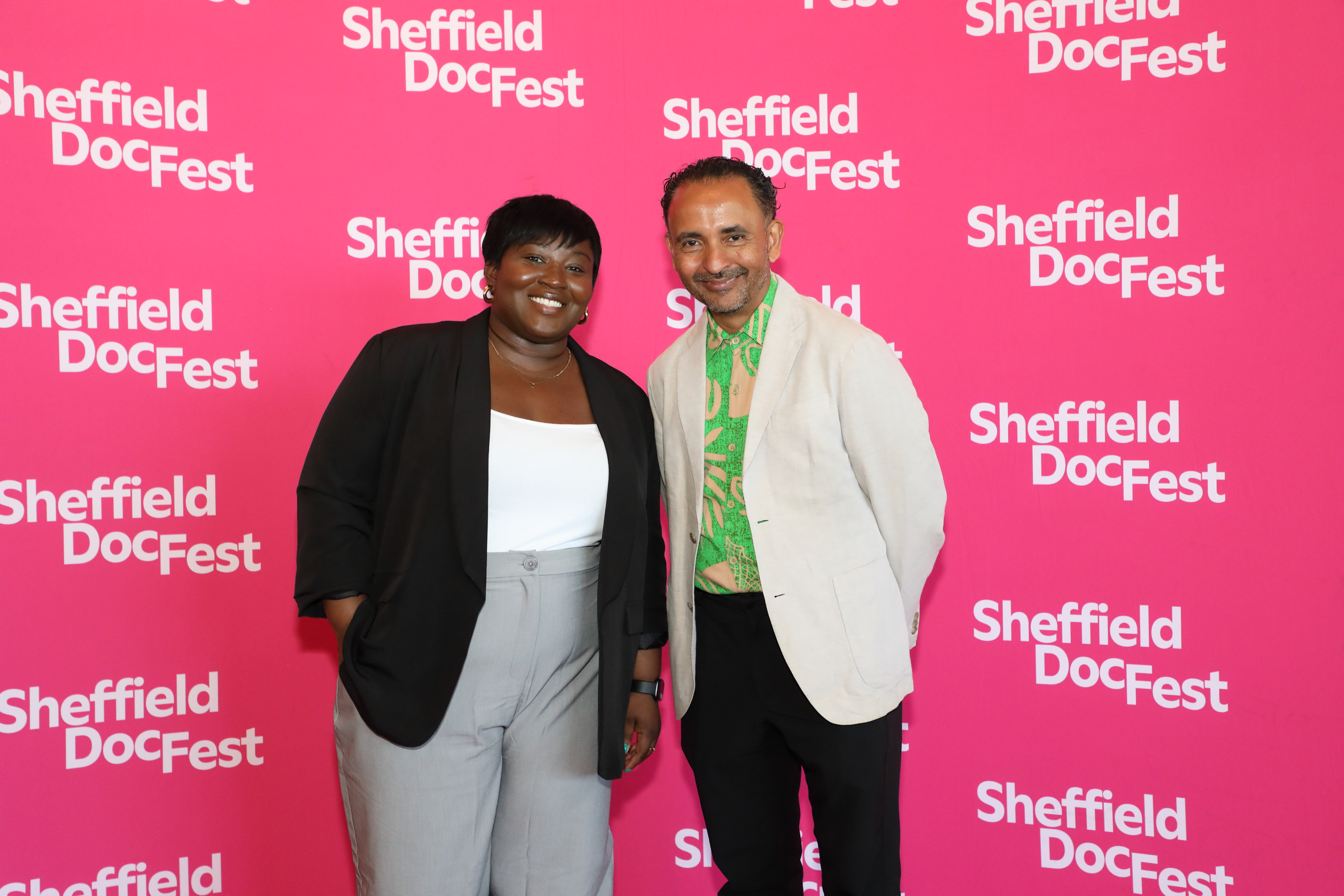 Grenfell: Uncovered director Olaide Sadiq, pictured with Festival Creative Director Raul Niño Zambrano smile as they stand against a bright pink and white branded backdropat Sheffield DocFest 2025