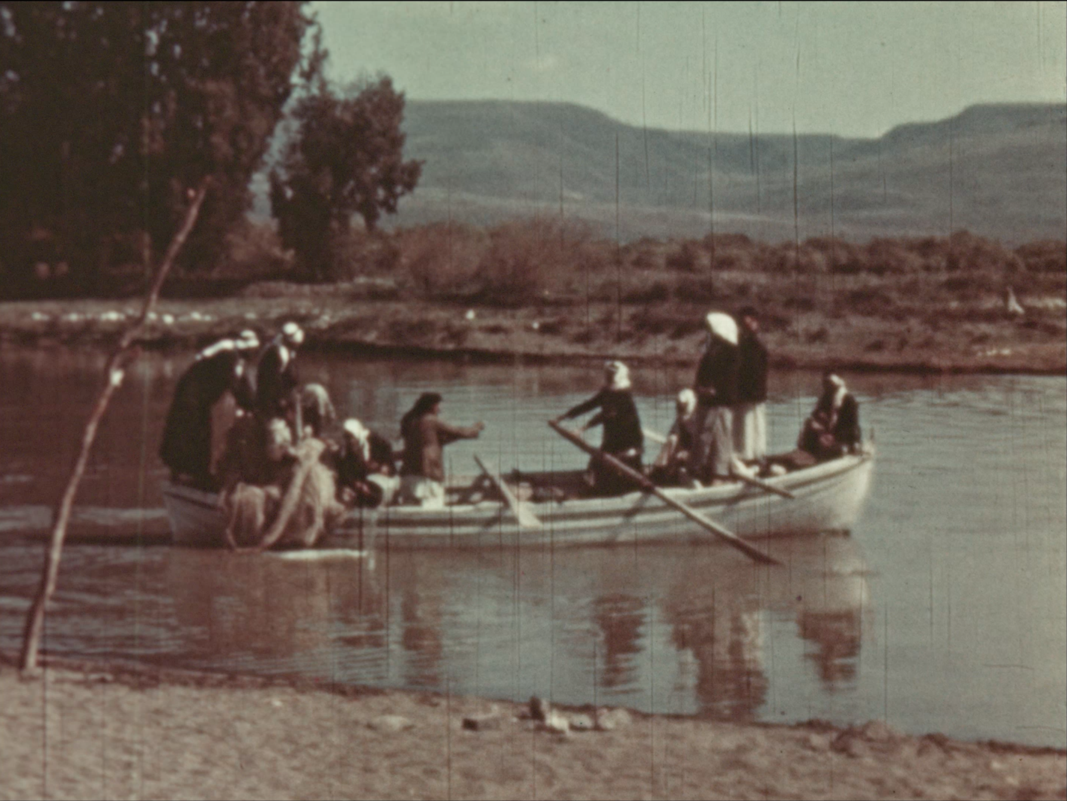 A group of people and animals in small boats crossing a calm river, with a rural landscape and hills in the background.