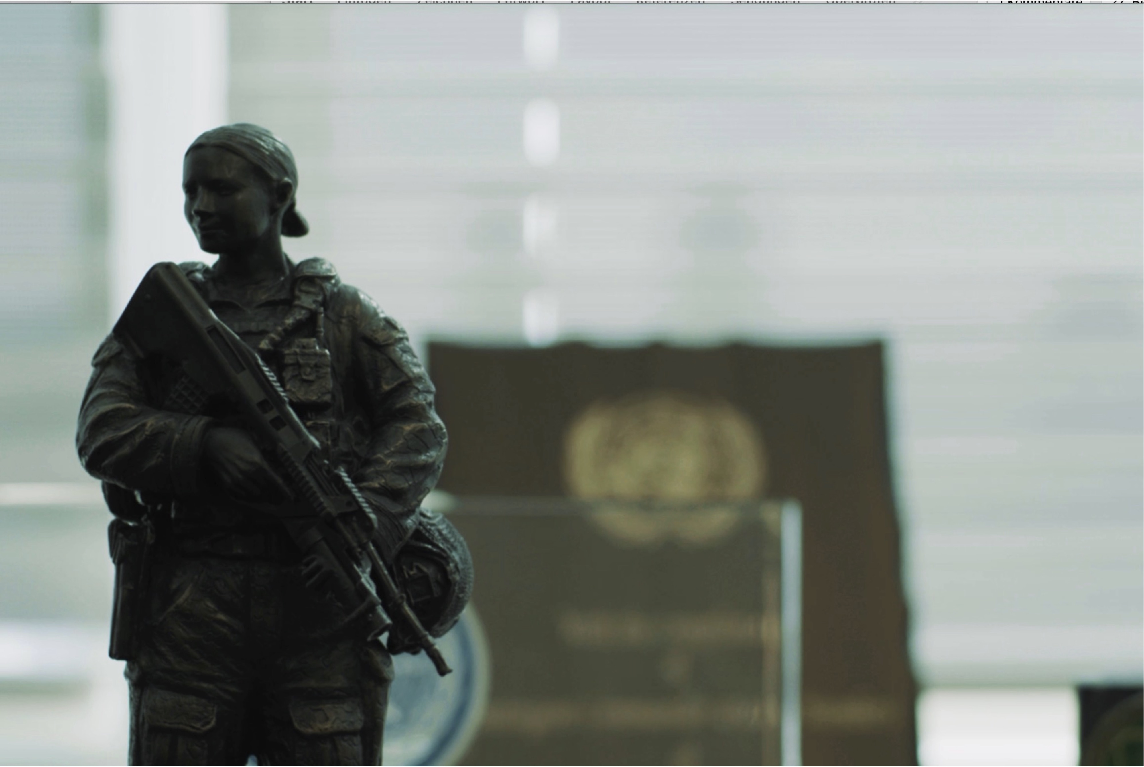 A small statue of a female soldier holding a rifle, with a blurred podium and emblem in the background, suggesting an official or institutional setting.