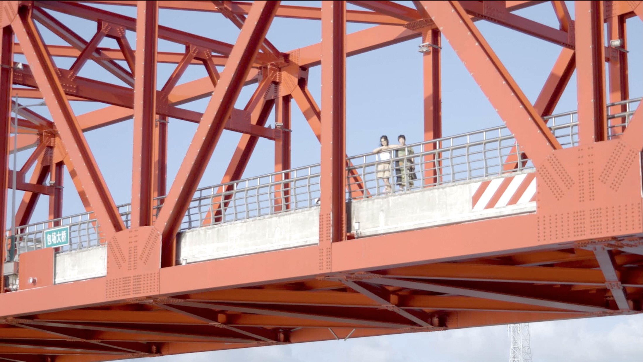 Two people stand side by side on a pedestrian walkway of a large red steel bridge, framed by its geometric beams, looking outward.
