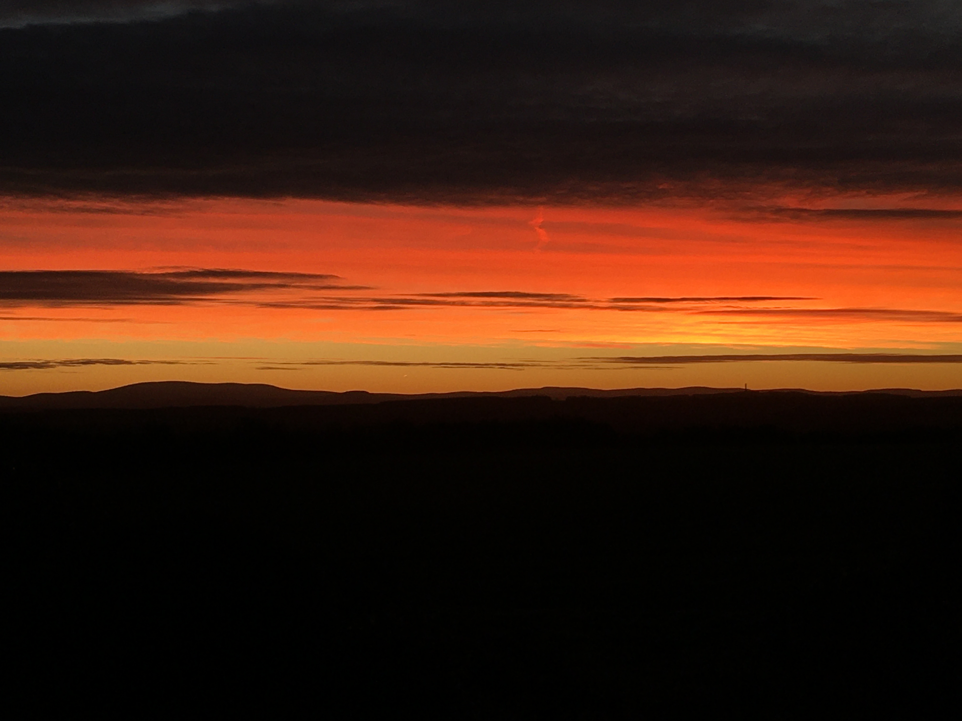 A bright orange-red sunset over dark, silhouetted hills with streaks of clouds above.