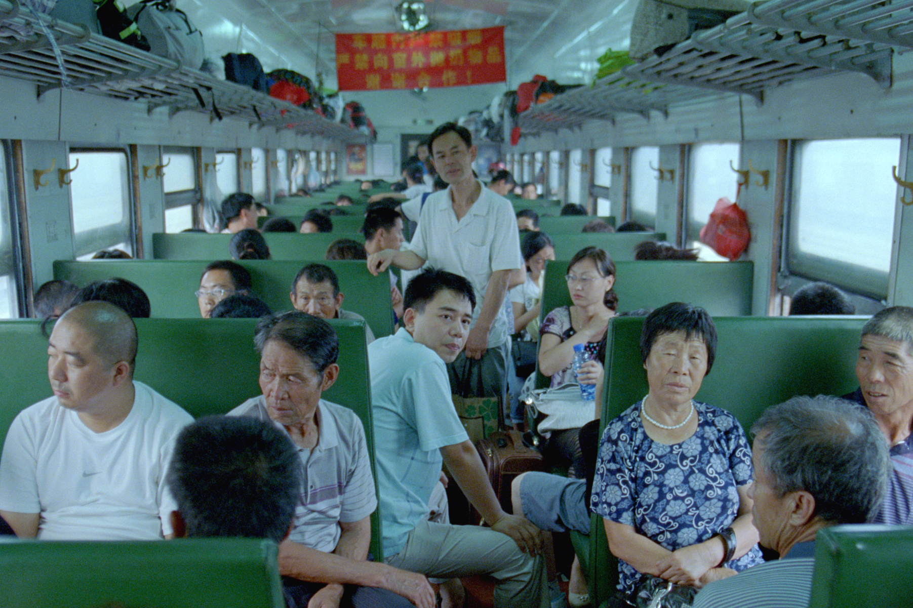 A crowded train carriage with rows of green seats filled with passengers. A man stands in the aisle while others sit facing each other, some looking toward the camera. 