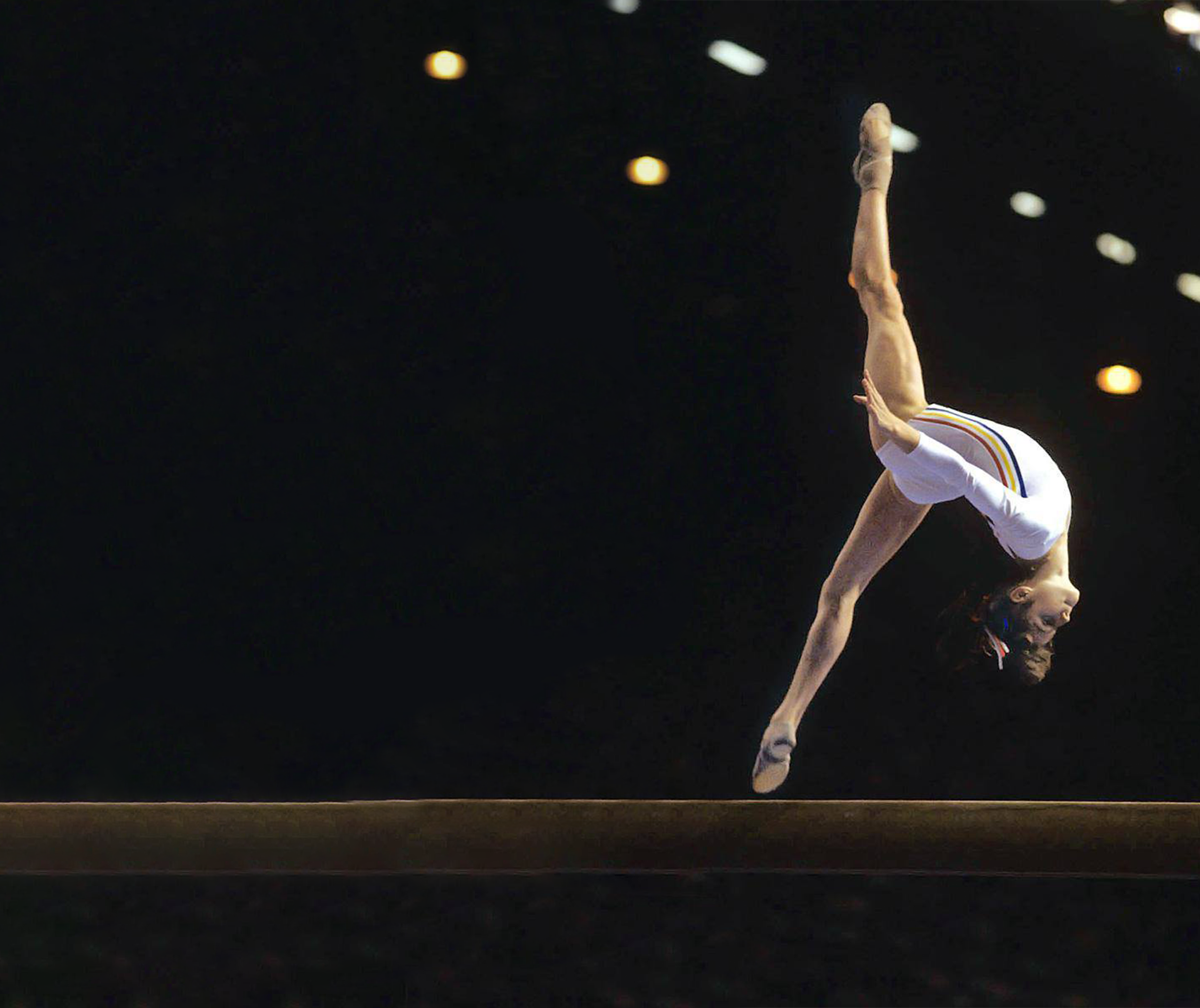 A gymnast in a white leotard performs a dynamic, arched split-leap mid-air above a balance beam against a dark, spotlighted arena background.