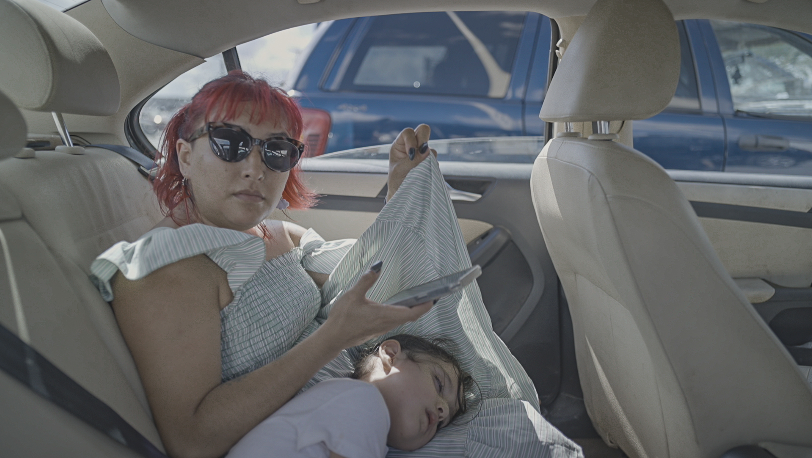 A woman with short red hair and sunglasses sits in the back seat of a car, holding a phone, while a young child sleeps on her lap.