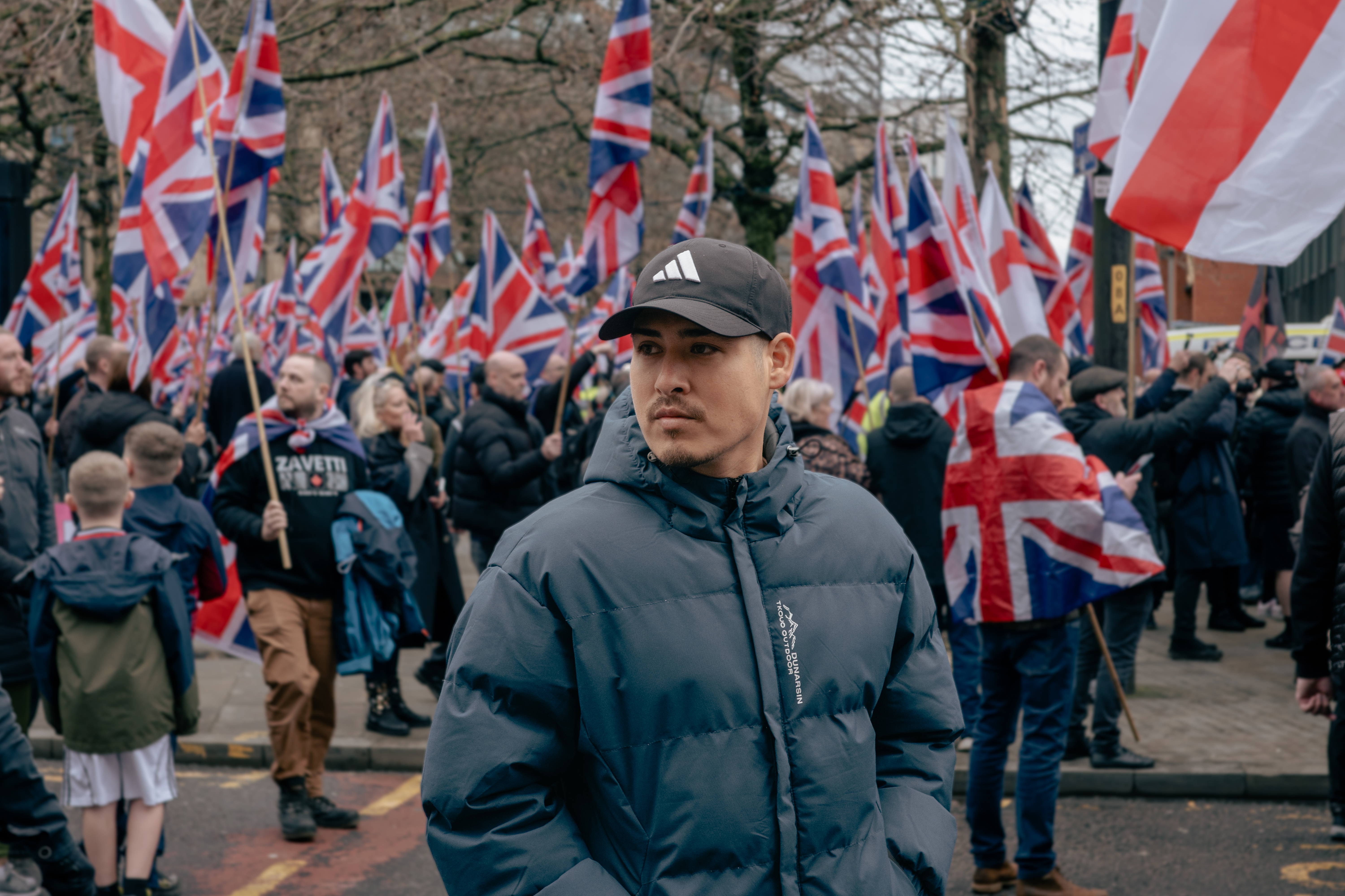 Anywhere but here  A man in a dark puffer jacket and cap stands in the foreground, looking to the side, while a large crowd behind him holds numerous Union Jack flags at a street protest.