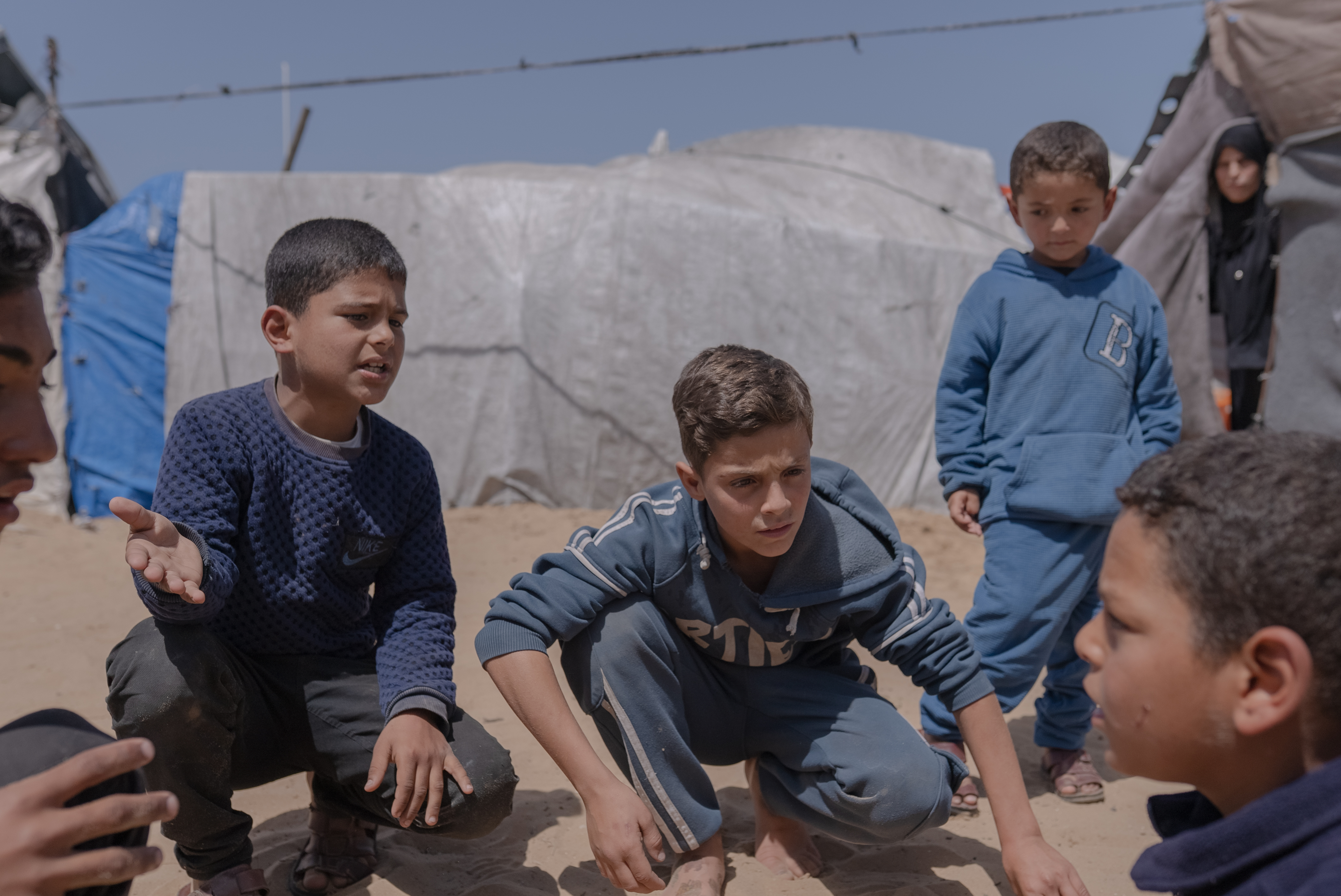 Several children gather and crouch in a sandy area between makeshift tents, appearing focused on a game or conversation. The scene takes place in a camp setting under a clear blue sky, with tarps and temporary shelters in the background.